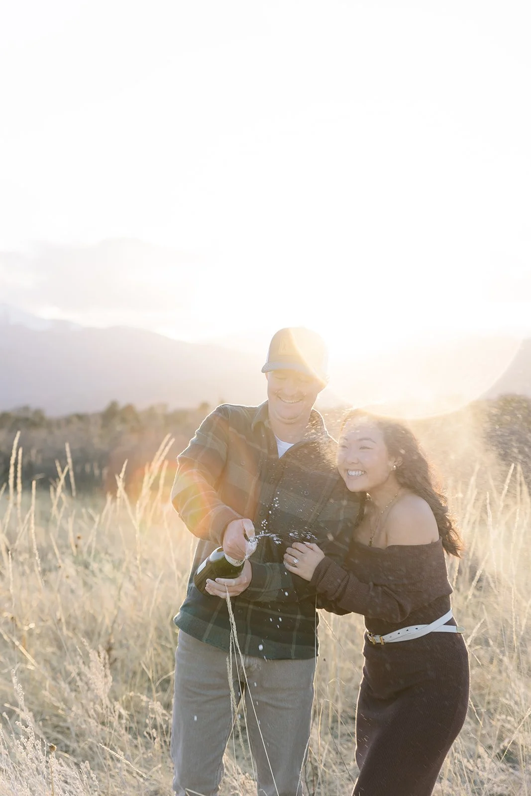 Two people celebrating outdoors at sunset, opening a bottle of champagne, with mountain landscape in background.