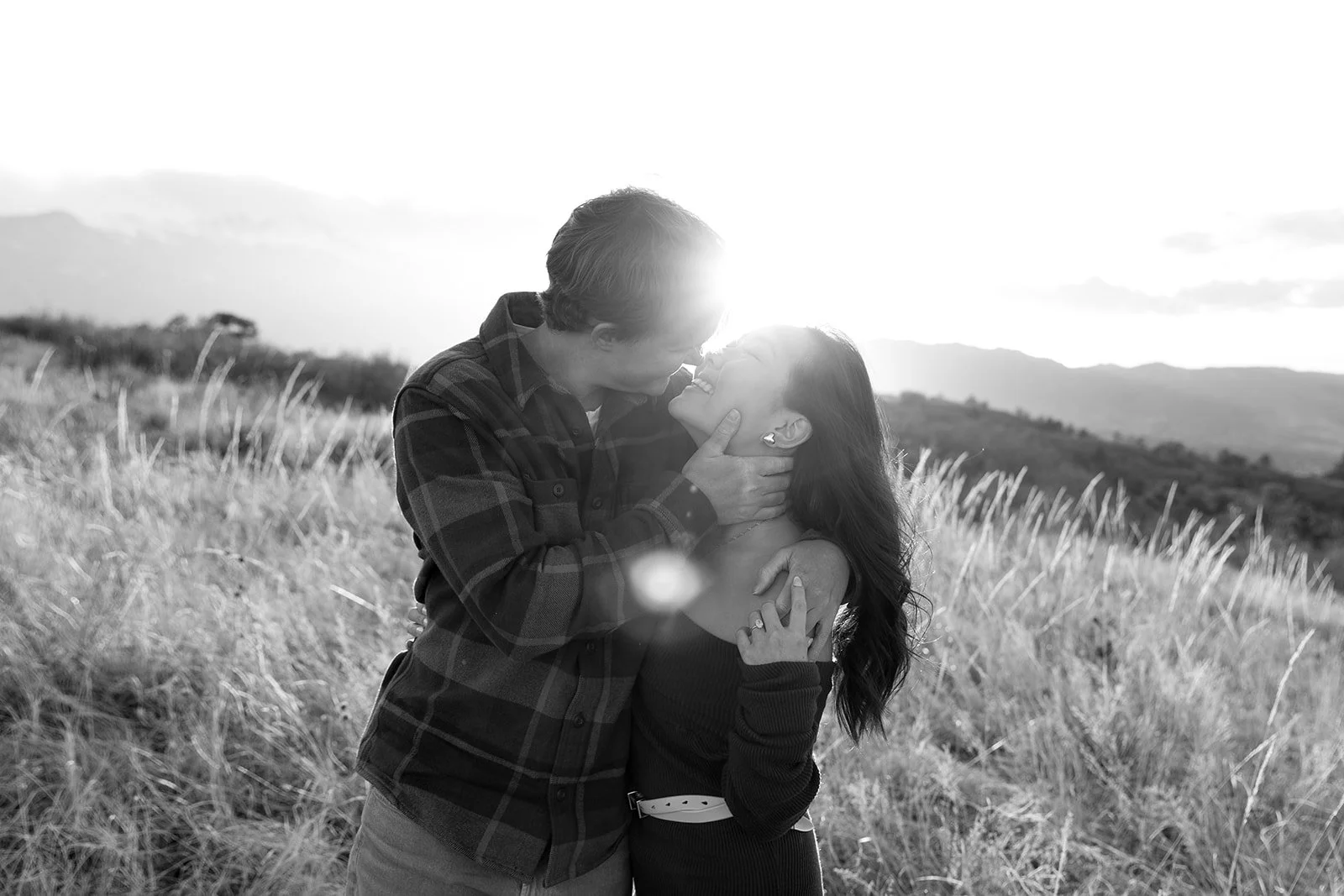 A black-and-white photo of a couple in a field, embracing and smiling as they lean in for a kiss, with the sun setting behind them.
