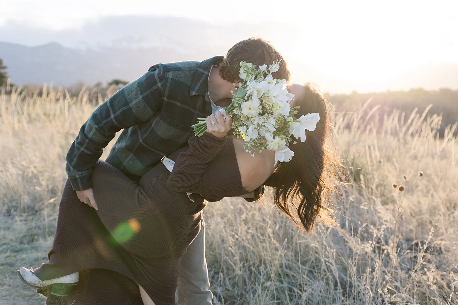 A couple is kissing outdoors during sunset, the woman is holding a bouquet of white flowers, and they are embracing in a field with tall dried grass, mountains in the background.