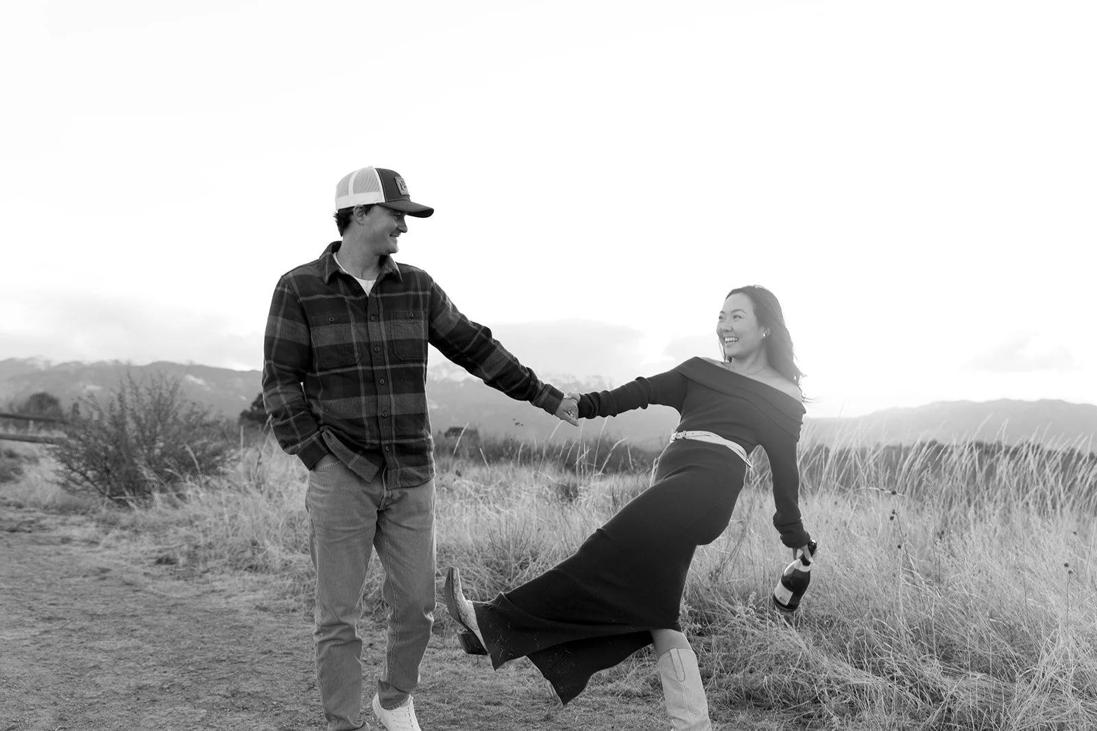 A young man and woman dancing outdoors in a field, holding hands and smiling, with mountains in the background, in black and white.