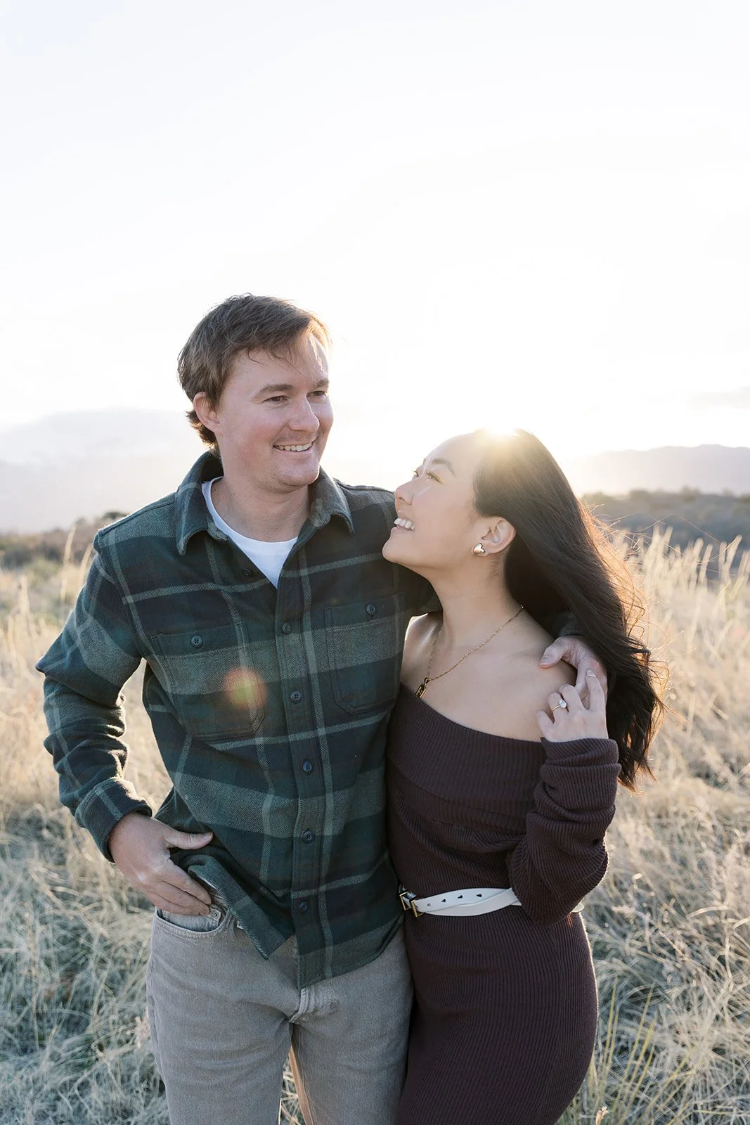 A smiling couple standing in a field with tall grass during sunset, looking lovingly at each other.