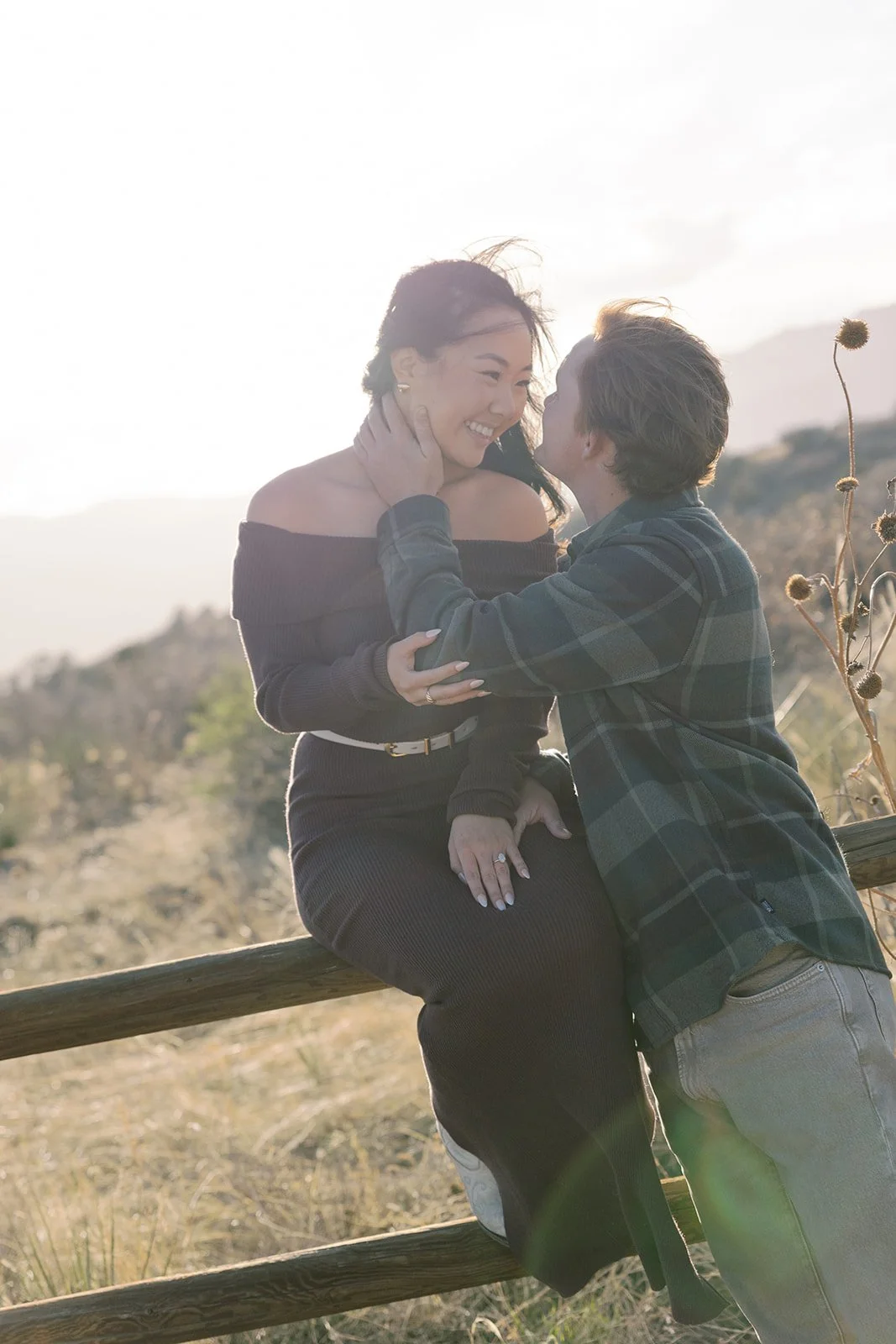 A couple sharing an intimate moment outdoors, with the woman sitting on a wooden fence and the man standing close, both smiling at each other in a natural setting.