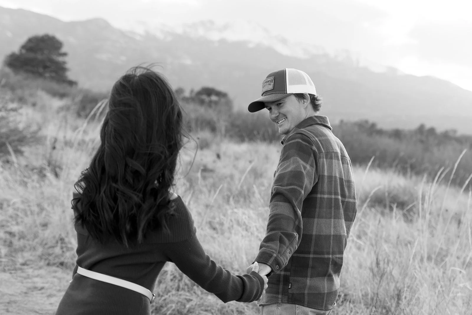 A man and woman holding hands and smiling at each other outdoors in a natural setting with mountains in the background.