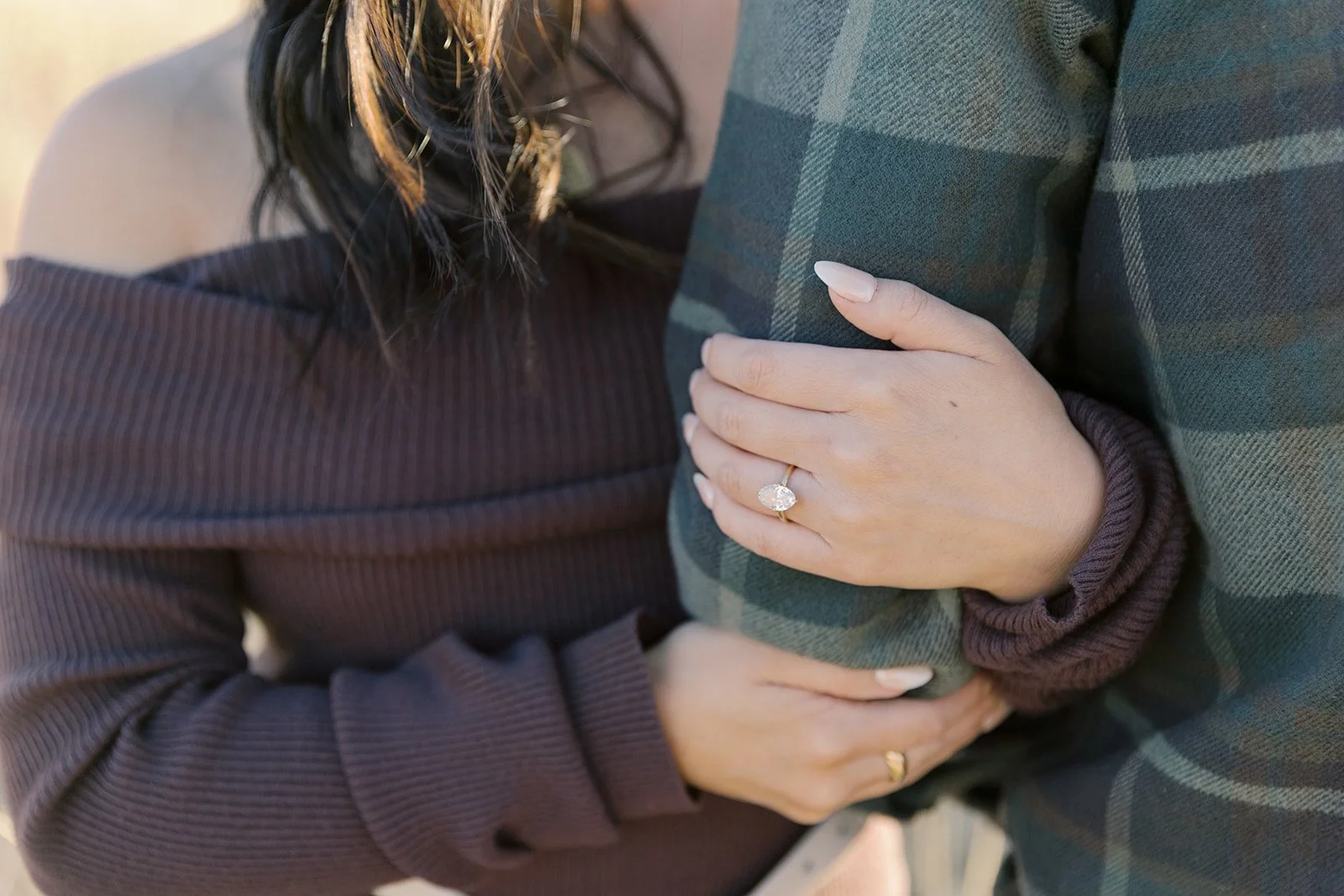A woman embracing a man, holding his arm with her hand, showing an engagement ring.