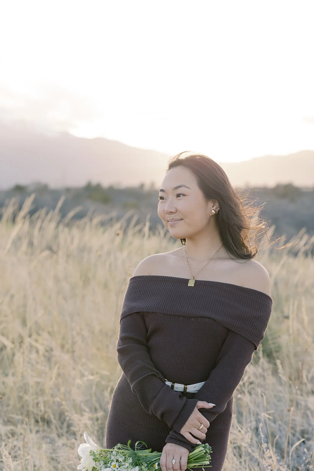 A woman in a black off-the-shoulder dress stands in a grassy field during sunset, holding a bouquet of white flowers.