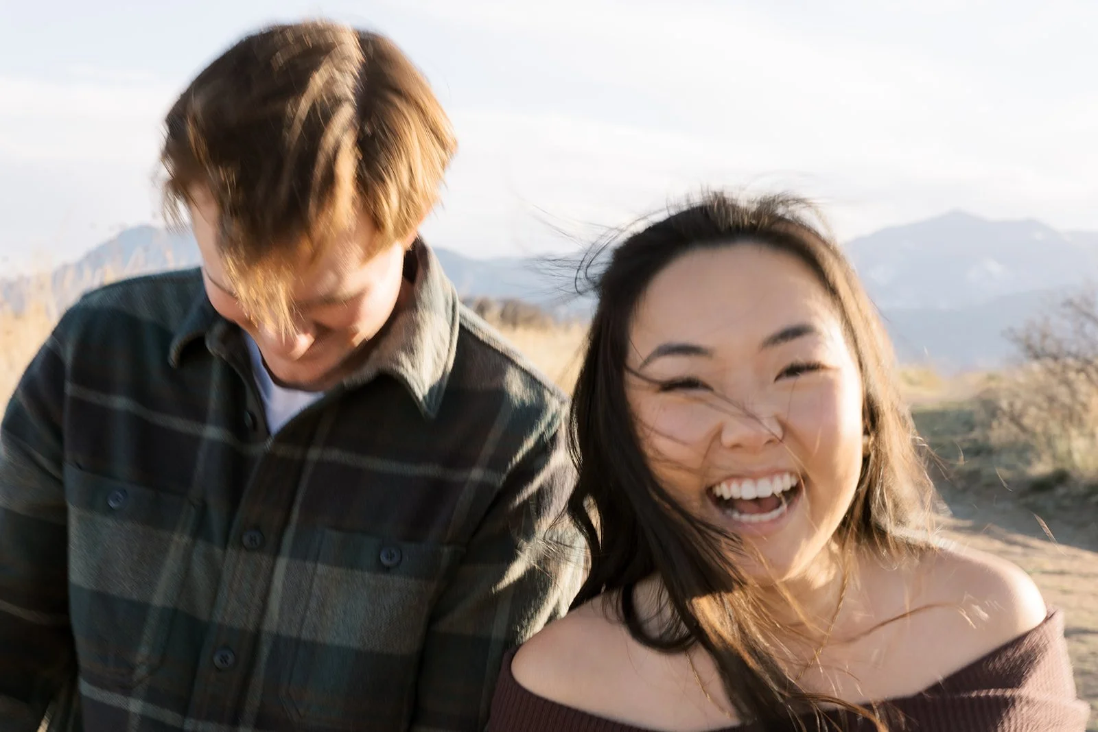 A young man and woman laughing outdoors in a natural landscape with mountains in the background.