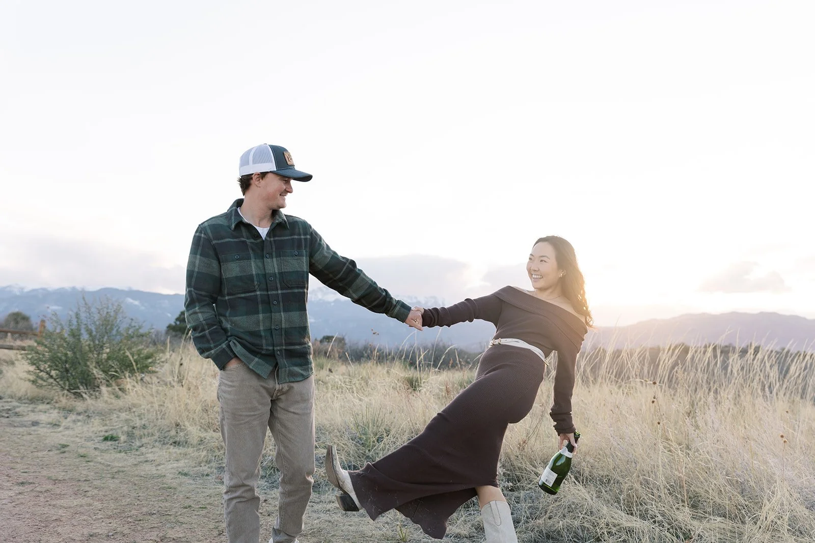 A young man and woman are outdoors in a field at sunset, holding hands and smiling. The woman is dressed in a black dress and boots, holding a bottle of champagne, while the man is dressed casually in a plaid shirt and baseball cap.