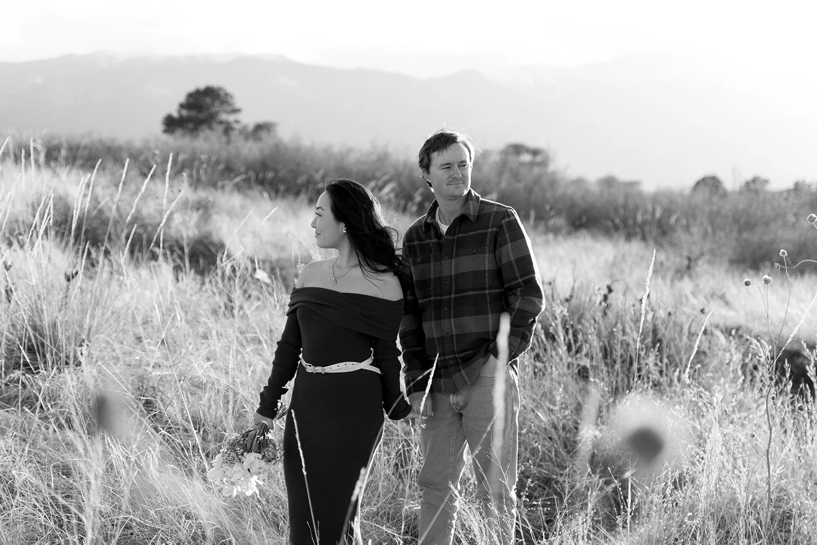 A man and woman walking through a grassy field holding hands, woman holding a bouquet, black and white