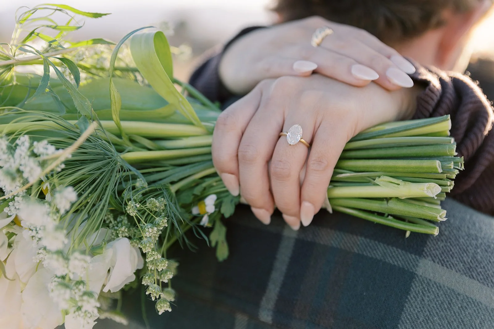 Close-up of a woman's hand with an engagement ring, resting on a bouquet of green and white flowers.