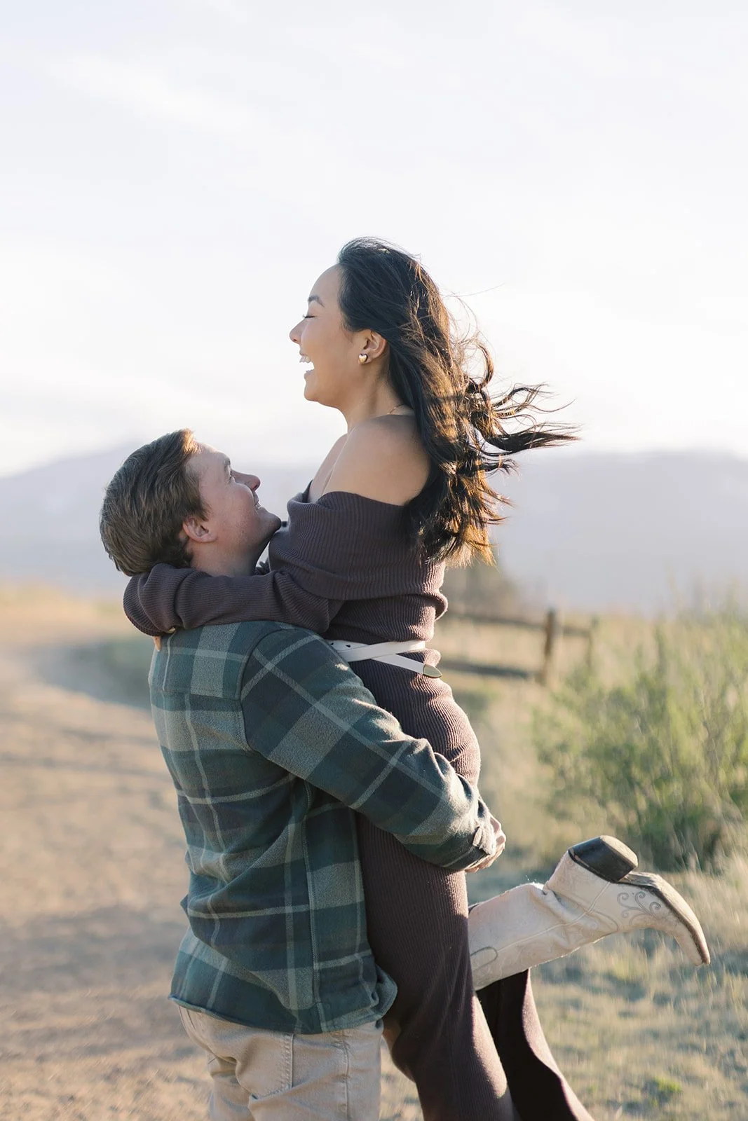A man is holding a woman in his arms outdoors, both smiling and laughing, with the woman having her arm around his neck. The scene appears to be during the daytime with a scenic background of a dirt path, grass, mountains, and a partly cloudy sky.