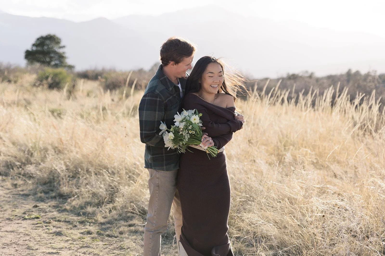 A man and woman standing in a field, smiling and embracing, the woman holding a bouquet of white flowers.