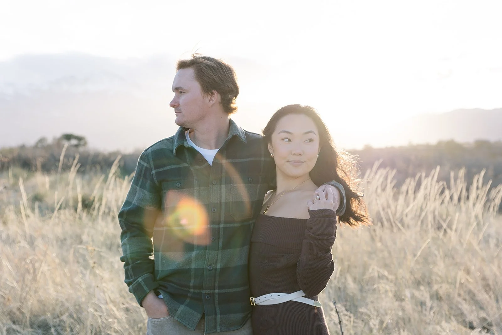 A couple standing in a field during sunset, with the man looking to the left and the woman smiling softly, holding his shoulder.