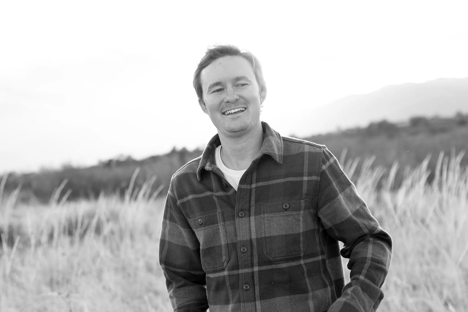 A smiling young man standing outdoors in a field with tall grass, wearing a plaid flannel shirt.