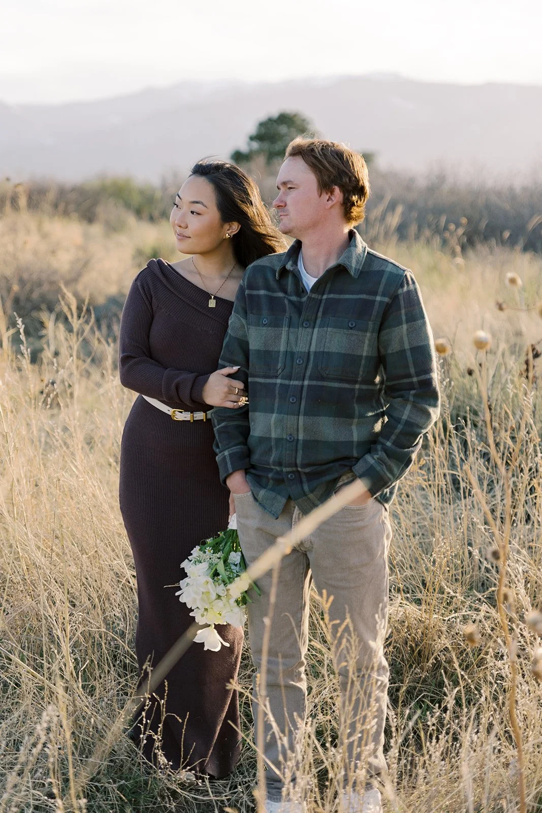 A couple standing in a field at sunset, with the woman holding a bouquet of white flowers, both appearing to be in a romantic or contemplative moment.