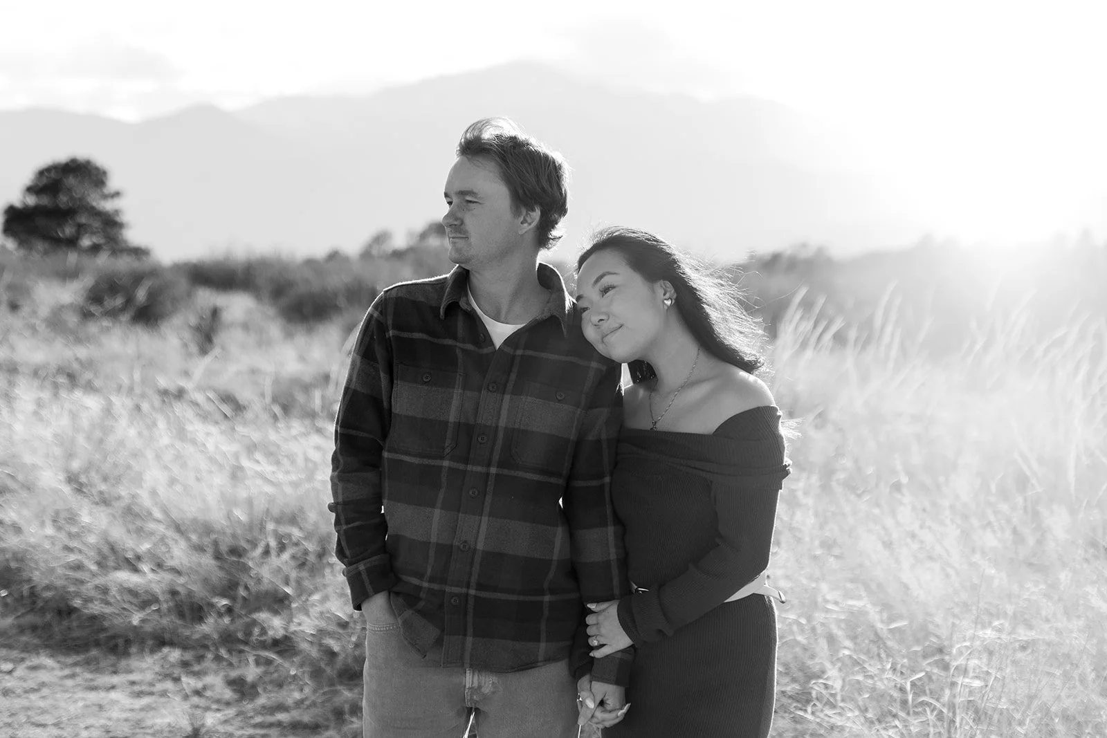 A black and white photo of a young man and woman standing outdoors in a field, holding hands. The woman rests her head on the man's shoulder, both smiling softly, with mountains and trees in the background.