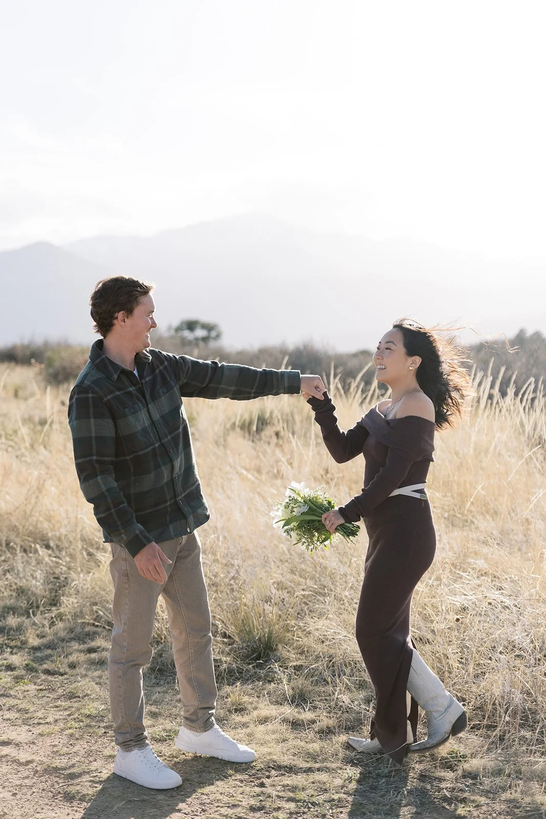 A man offers his fist to a smiling woman holding a bouquet of white flowers in a field of tall grass, with mountains in the background.