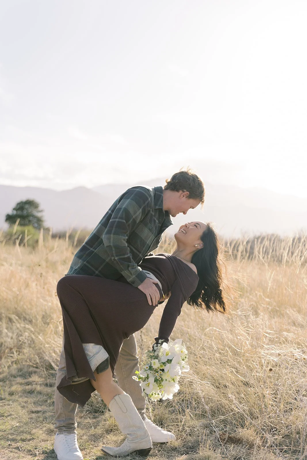 A couple enjoying a romantic moment in a field during sunset, with the man leaning over the woman who is holding a bouquet of white flowers.