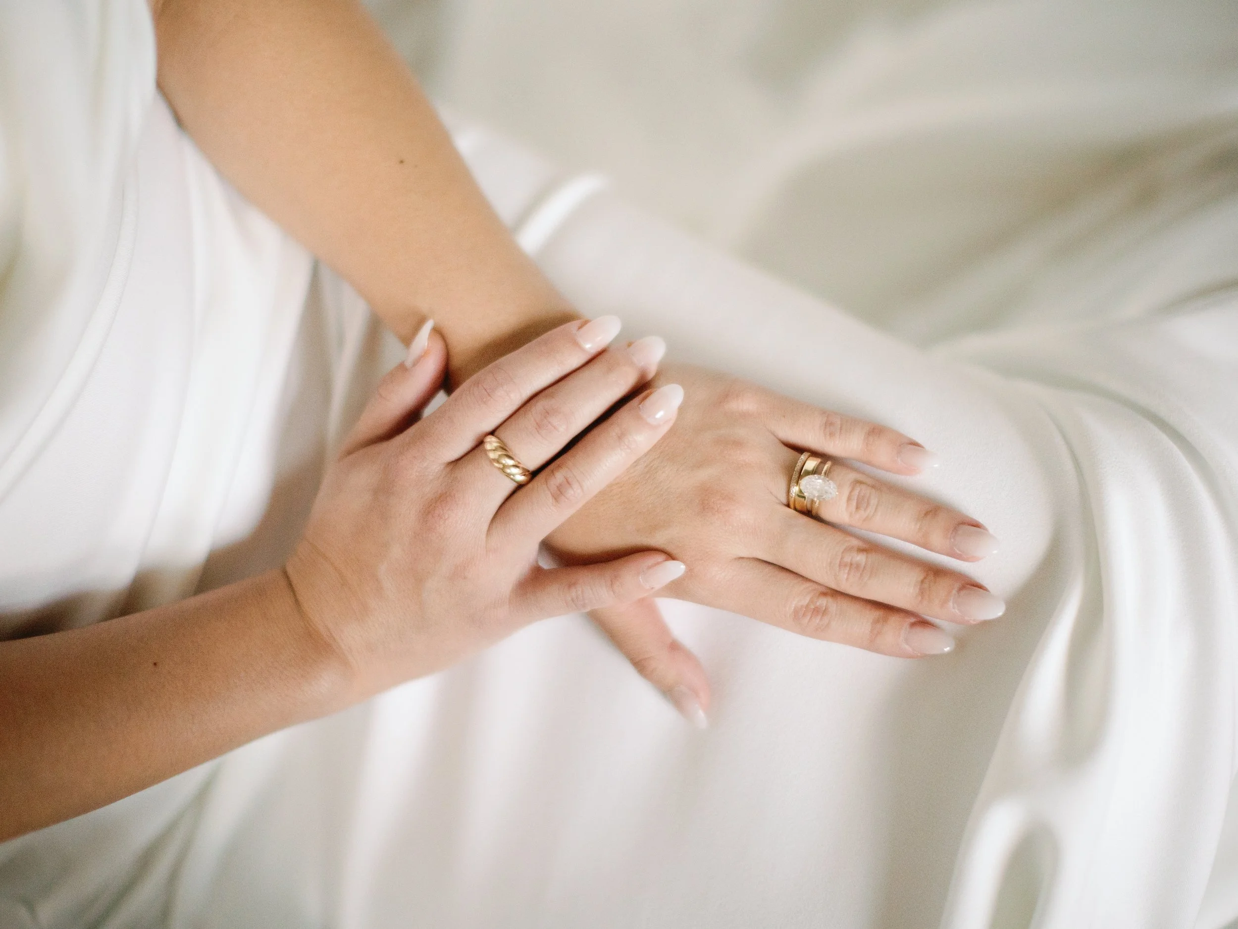 Close-up of a woman's hands resting on a silky white fabric, wearing multiple gold rings, one with a large diamond.