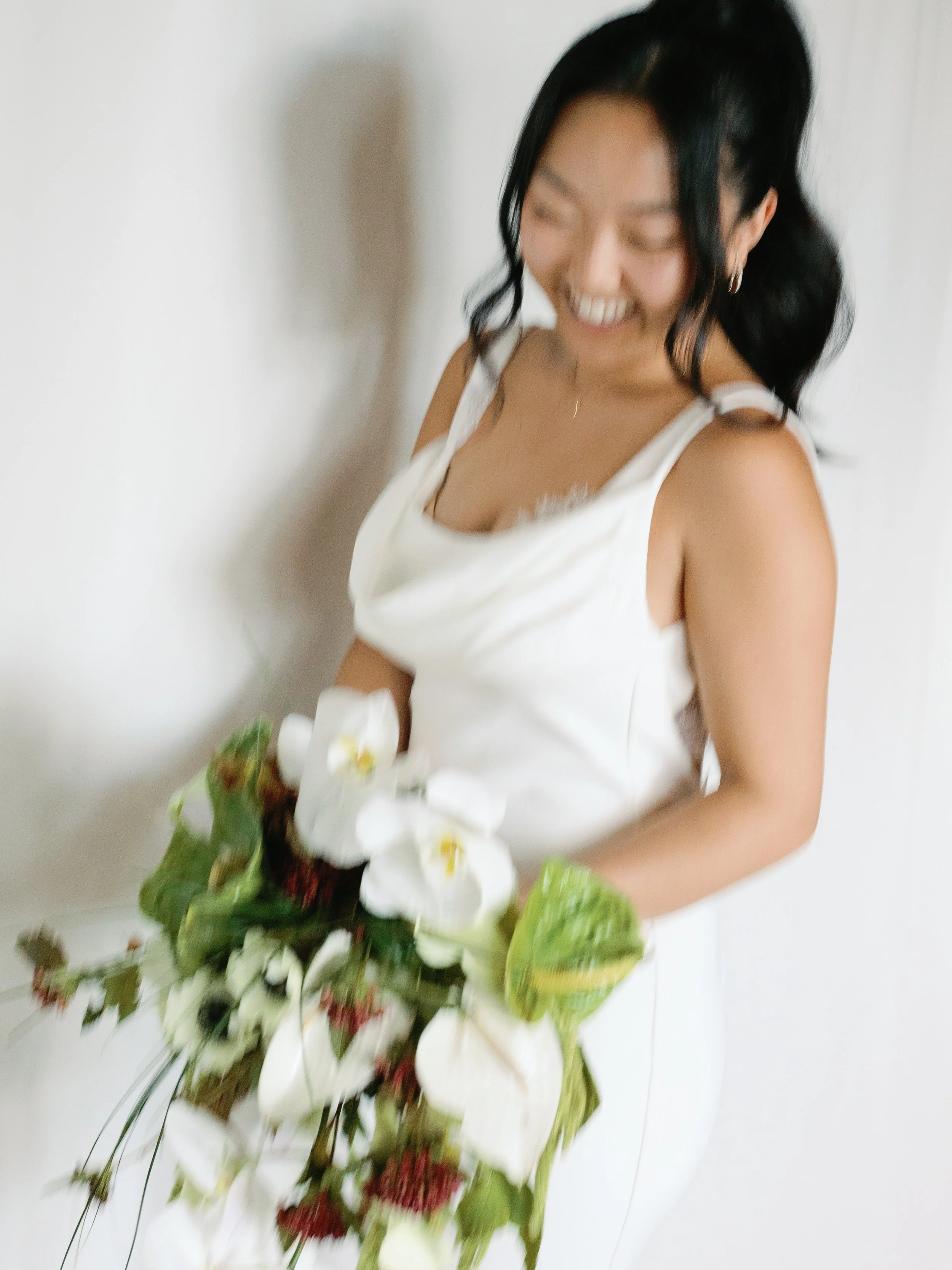 A woman in a white dress holding a flower bouquet, smiling and looking down.