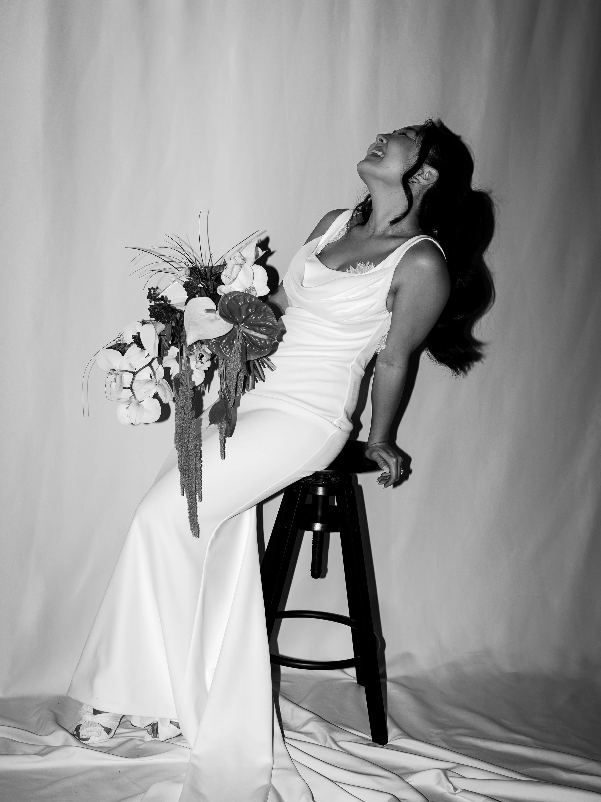 A woman in a white dress sitting on a stool, holding a bouquet of flowers, laughing with her head tilted back.