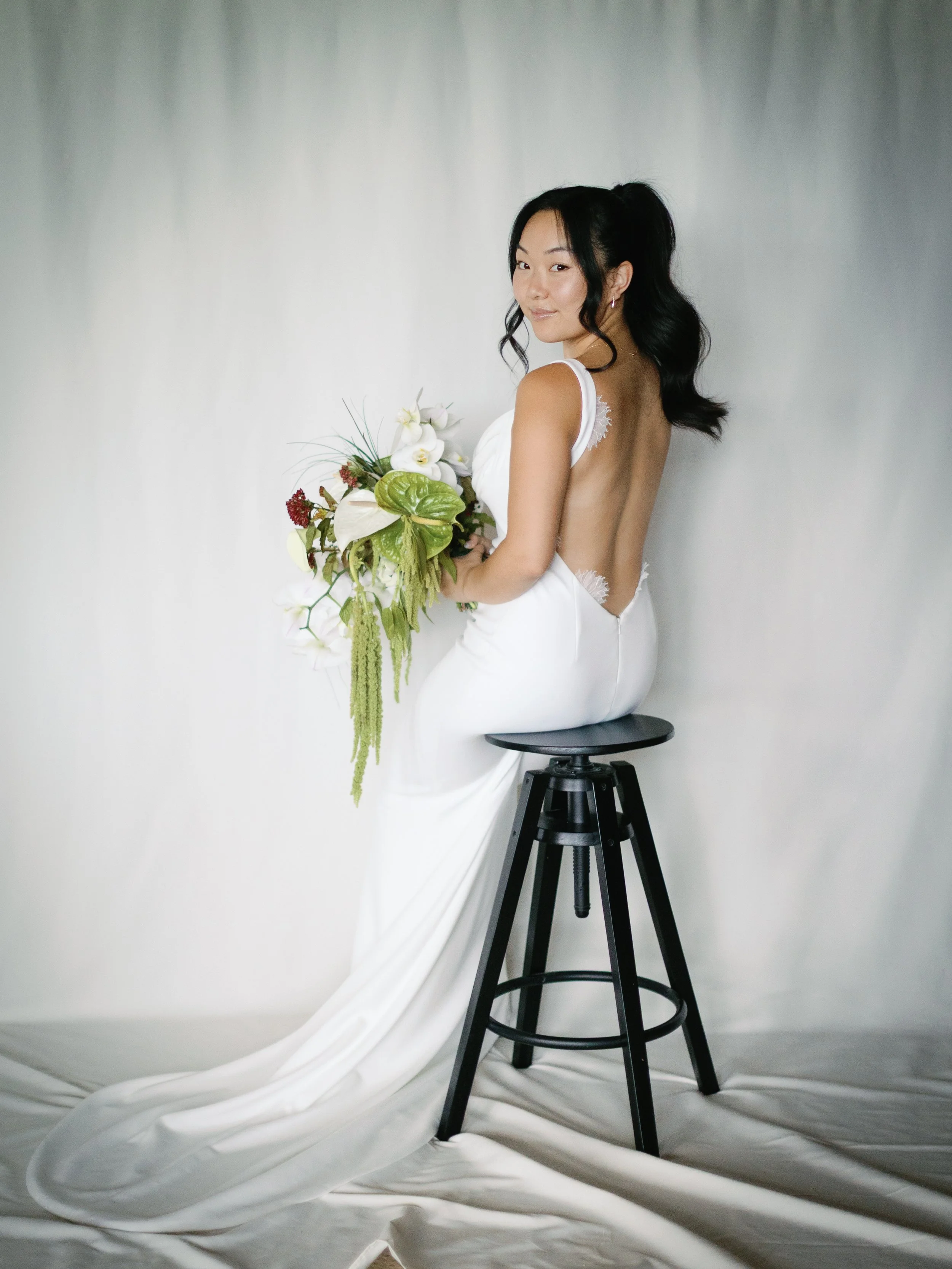 Bridal portrait of a woman in a white gown sitting on a black stool, holding a floral bouquet, with a plain light-colored background.