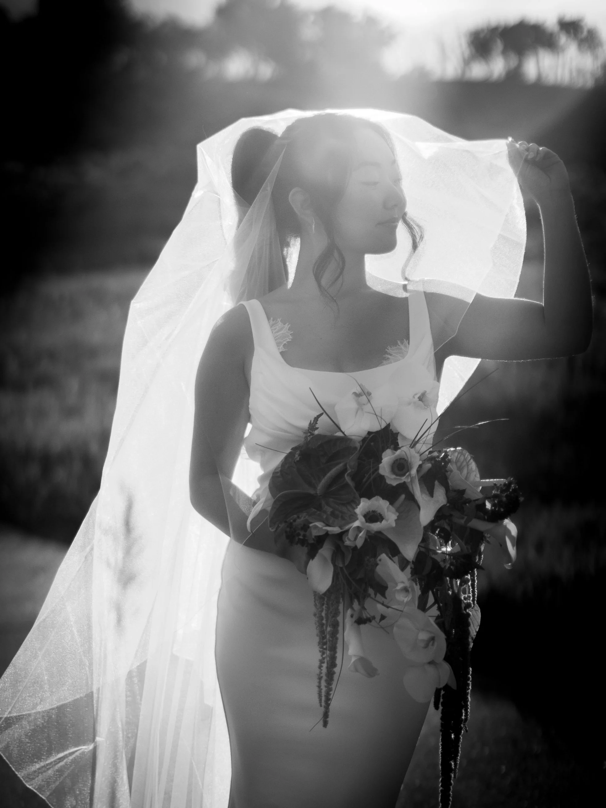 A woman in a wedding dress holding a bouquet of flowers, standing outdoors in sunlight with a veil over her face.