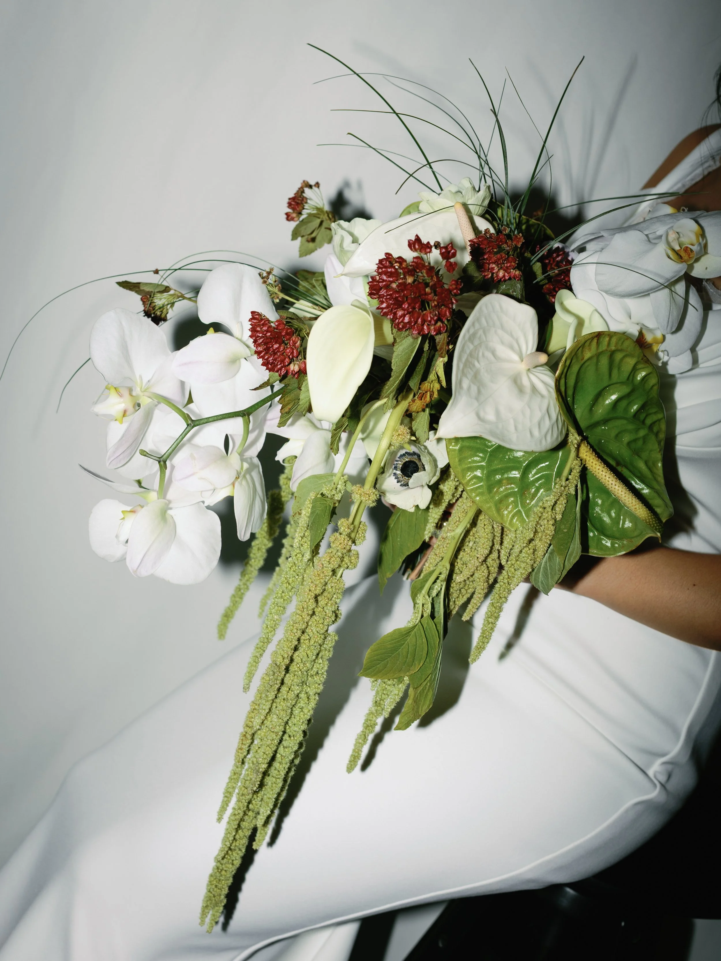 A person holding a bouquet of white calla lilies, white orchids, red flowers, and various green leaves and long green hanging amaranthus.
