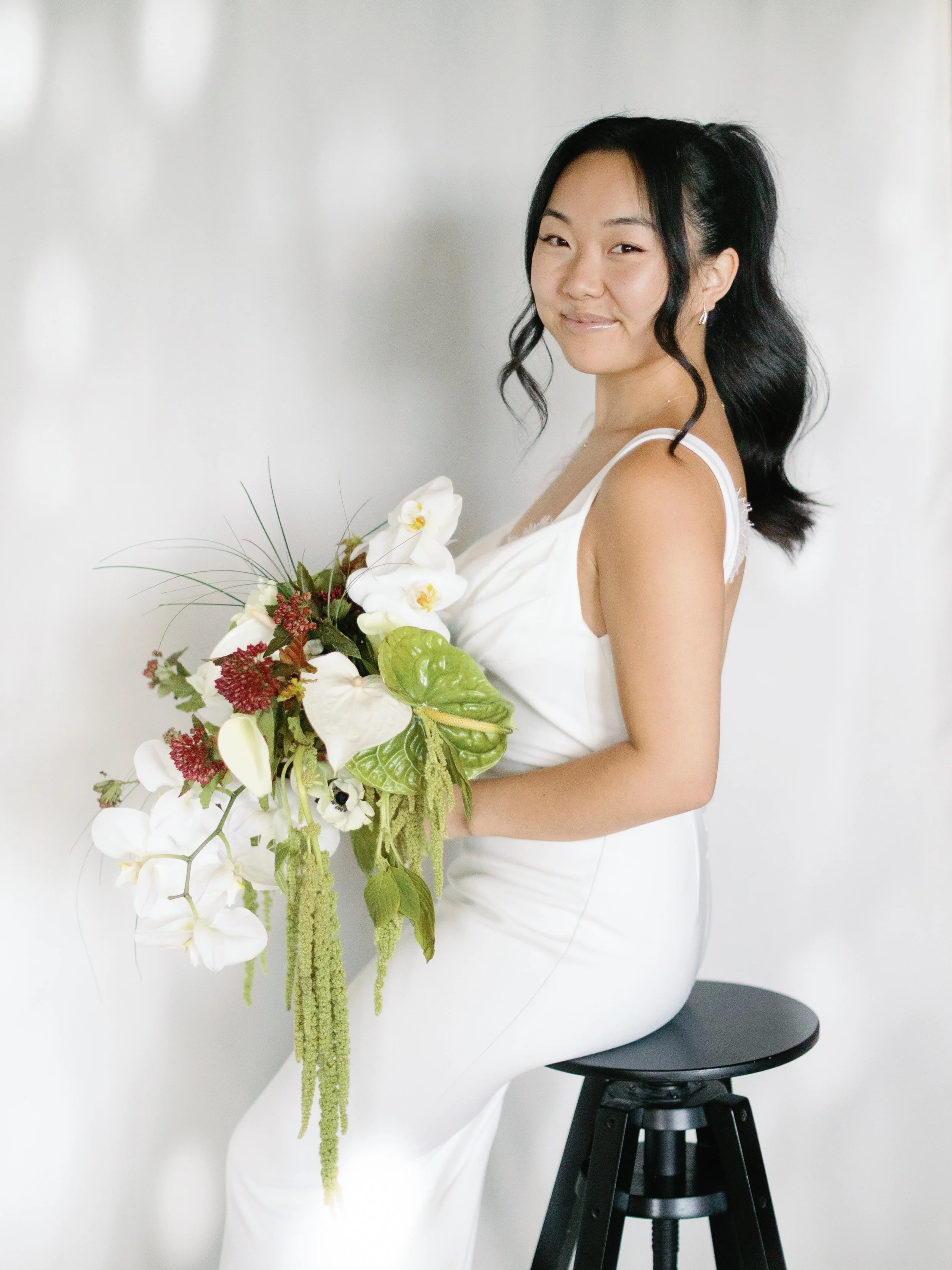 Young woman in white dress sitting on a black stool, holding a large bouquet of white and green flowers, with a slight smile, standing against a plain white background.