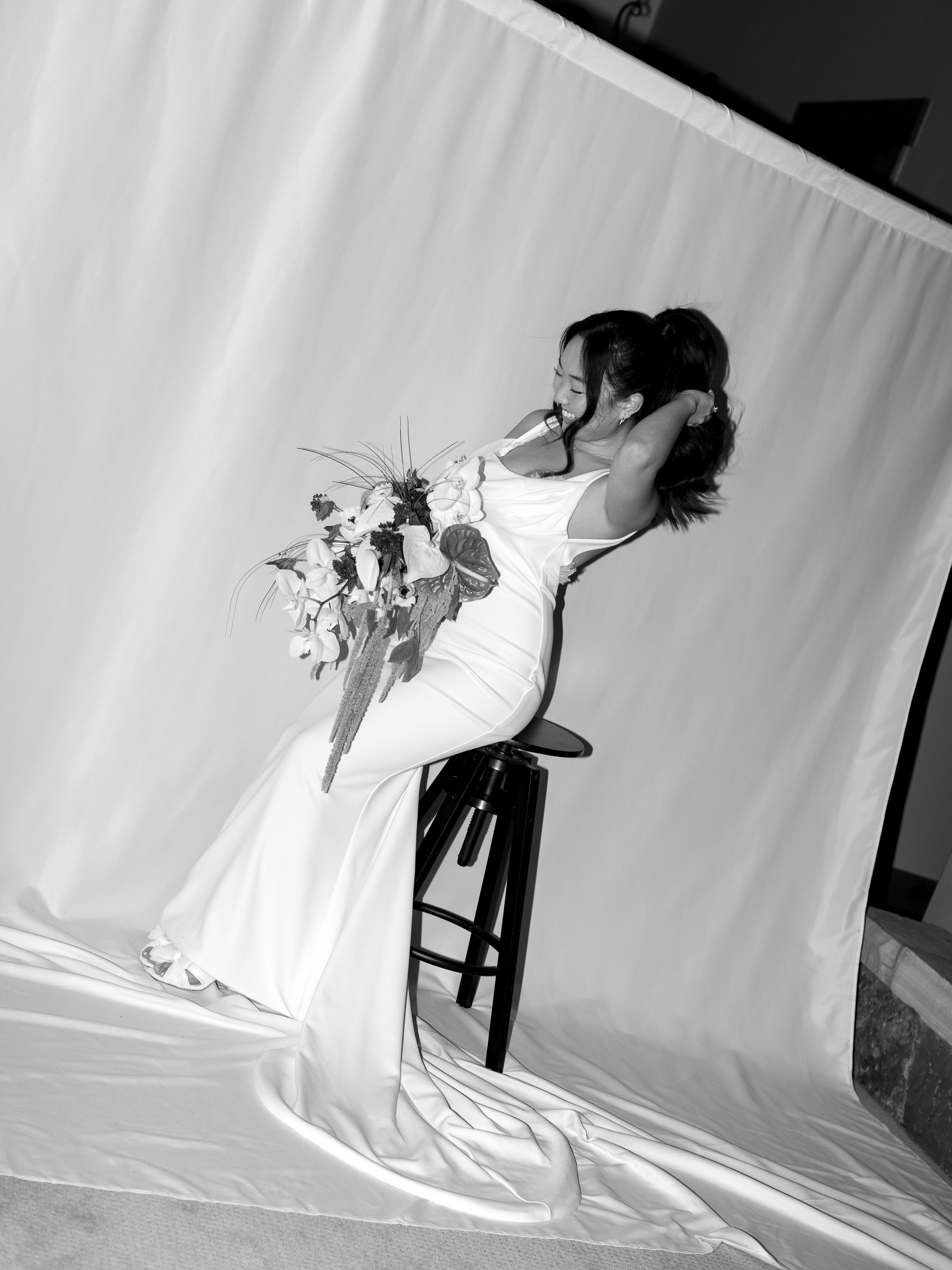 A woman in a white gown sitting on a stool, holding a large bouquet of flowers, smiling and laughing while tilting her head back.