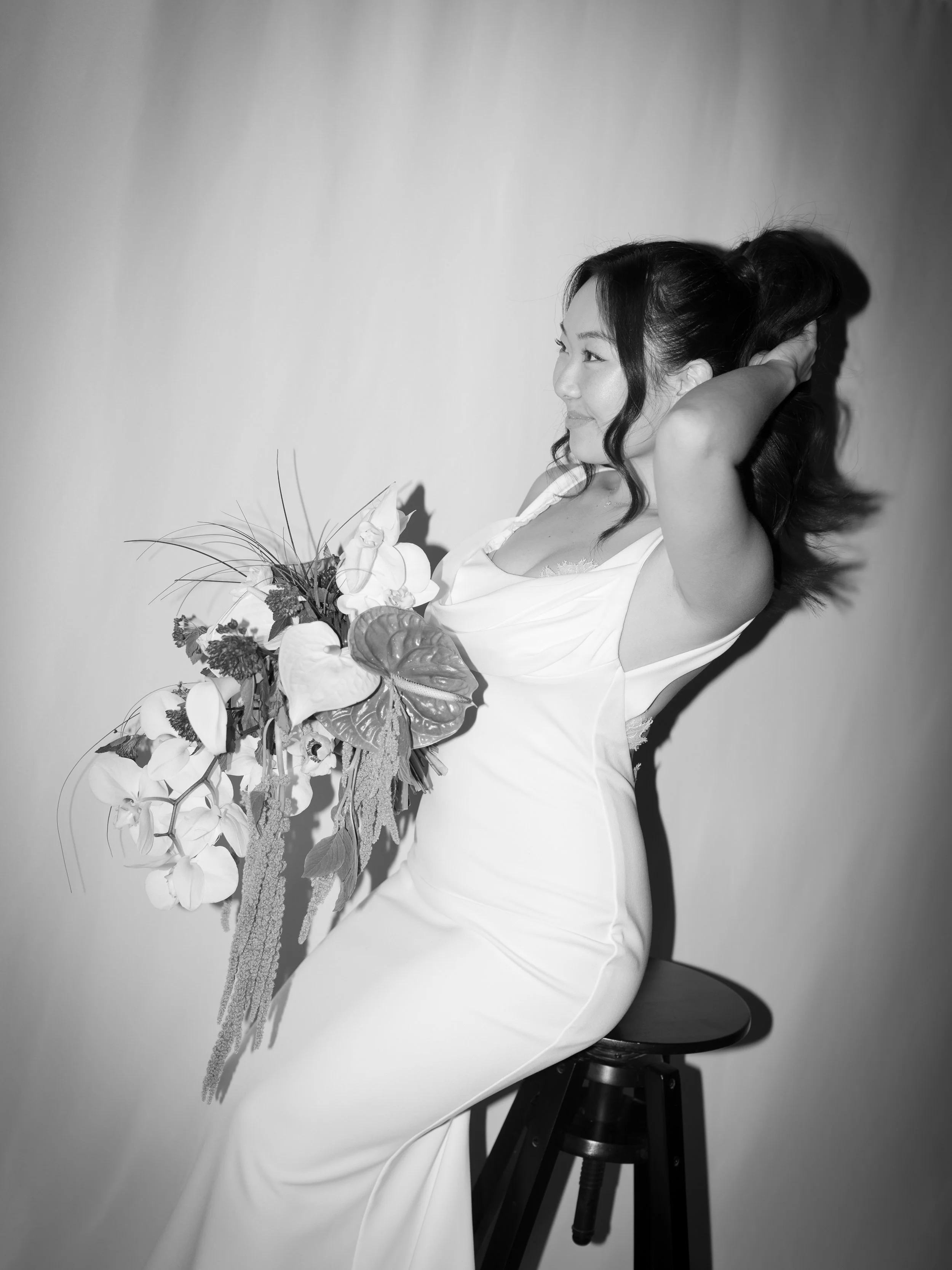A woman in a white dress sitting on a stool with a bouquet of flowers, touching her hair, smiling, against a plain background.