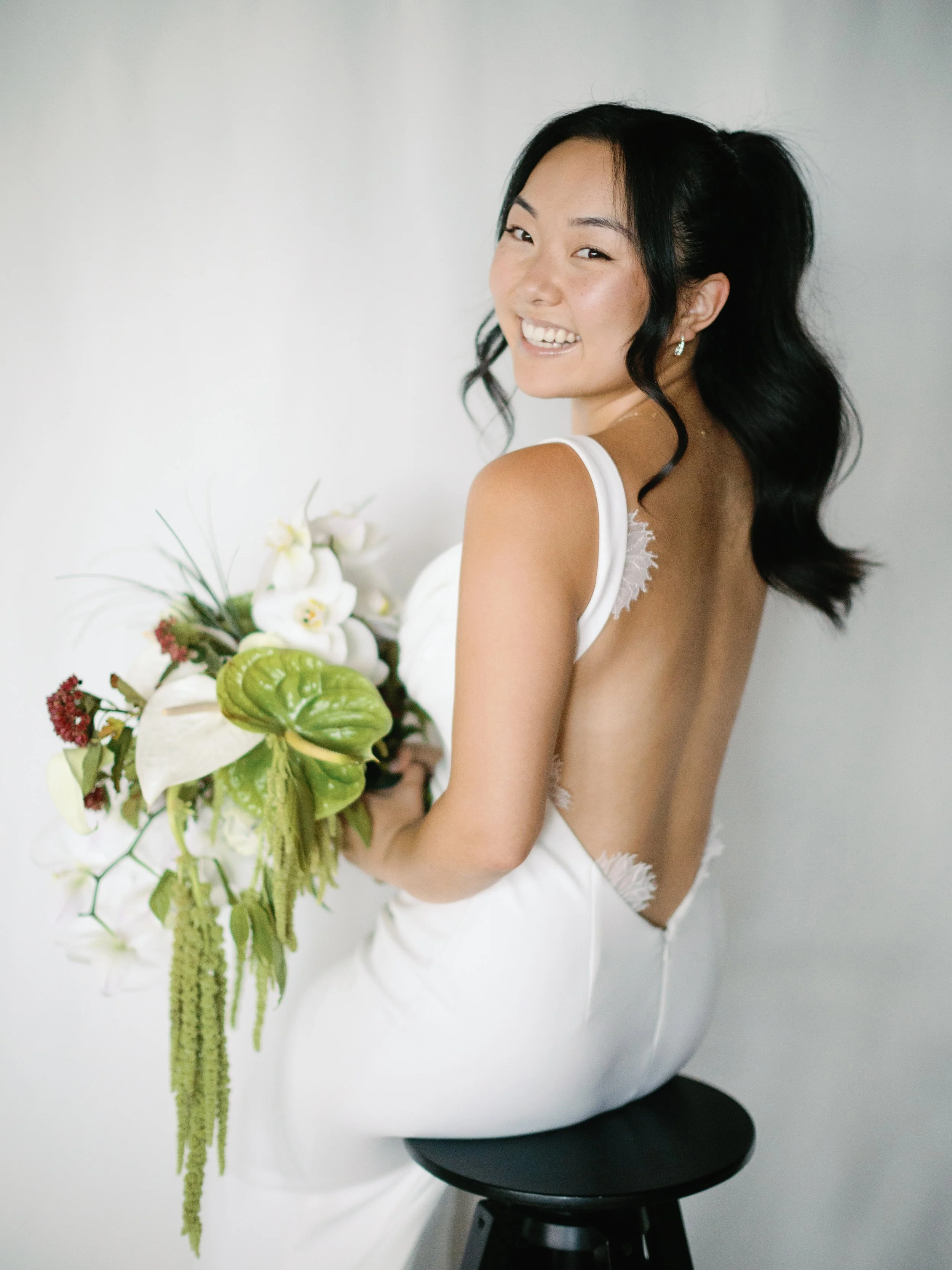 A smiling woman with black wavy hair in a ponytail, wearing a white dress with a deep back, sitting on a black stool and holding a bouquet of white and green flowers, against a plain light background.