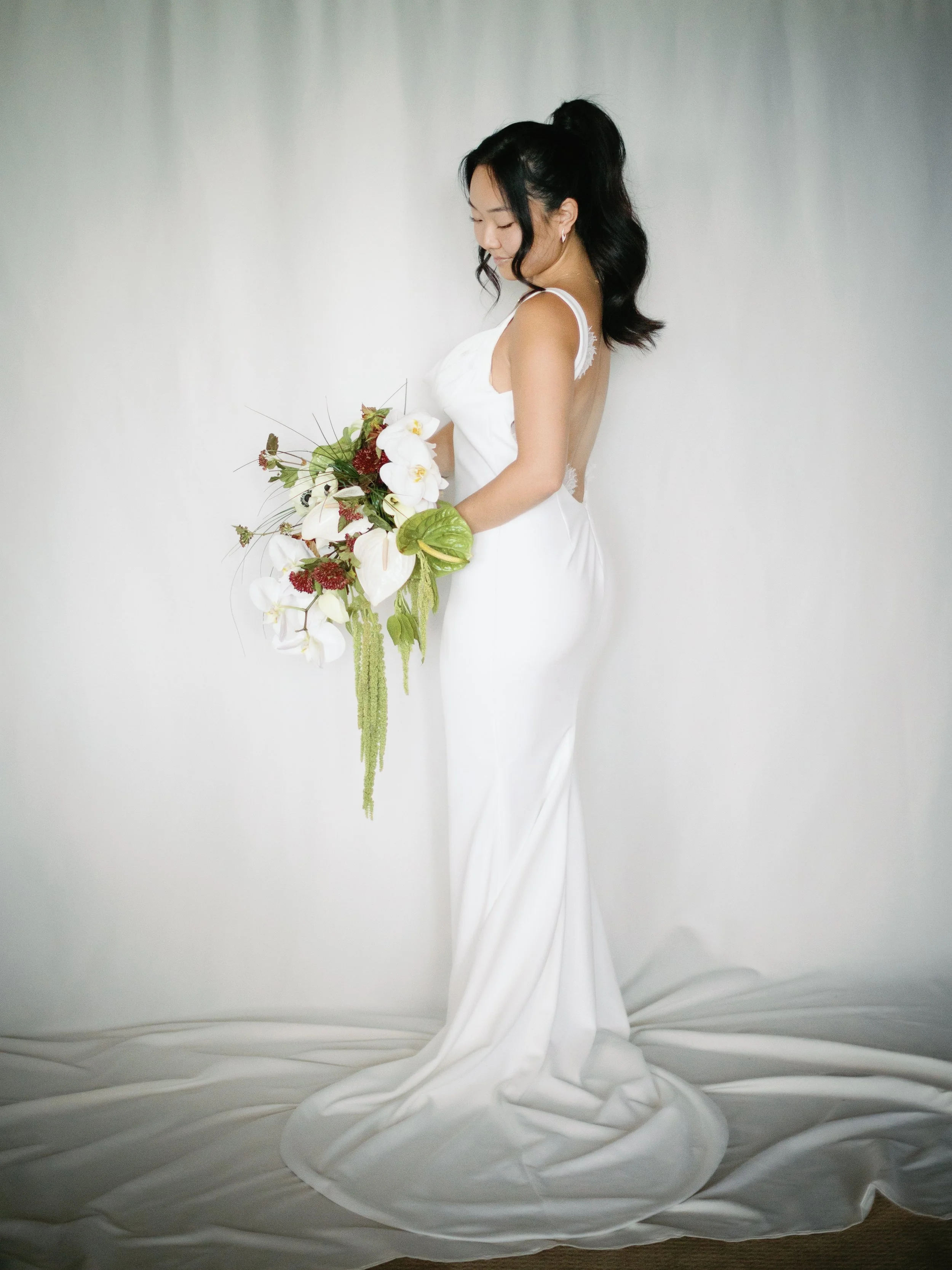 Woman in a white wedding dress holding a bouquet of white orchids and greenery, standing against a plain background.