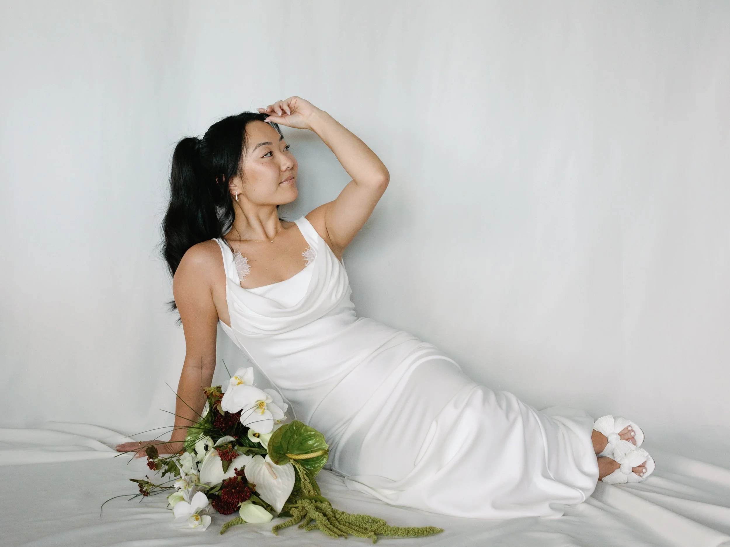 A woman in a white dress sitting on a white surface, holding a bouquet of white and green flowers, with her other hand resting on the surface, and looking to her right against a plain white wall.