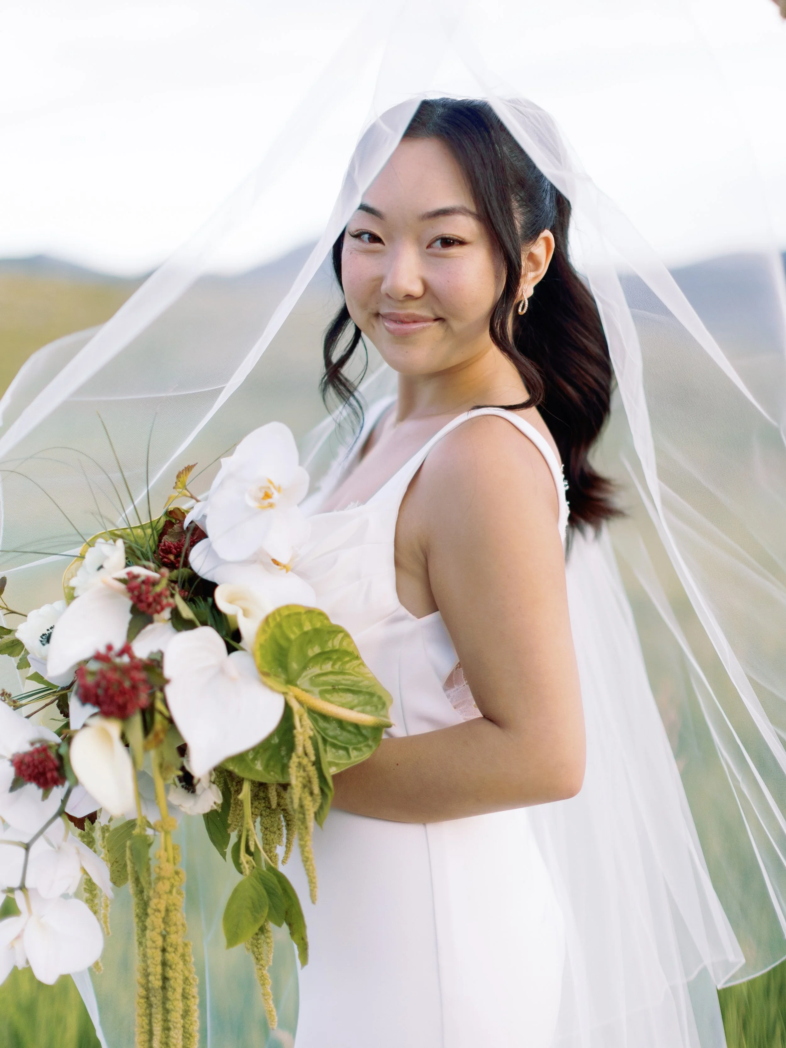 A smiling bride holding a bouquet of white flowers, wearing a white wedding dress and veil outdoors with a green landscape background.