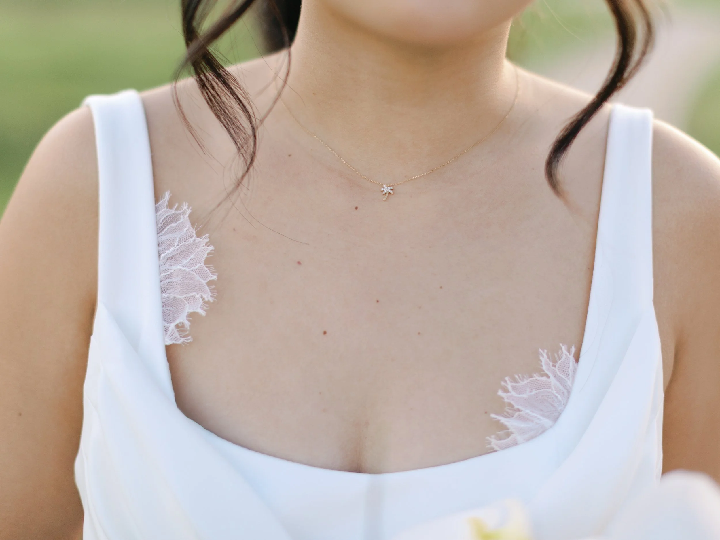 Close-up of a woman wearing a white dress with lace details on the straps and a delicate gold necklace with a small charm