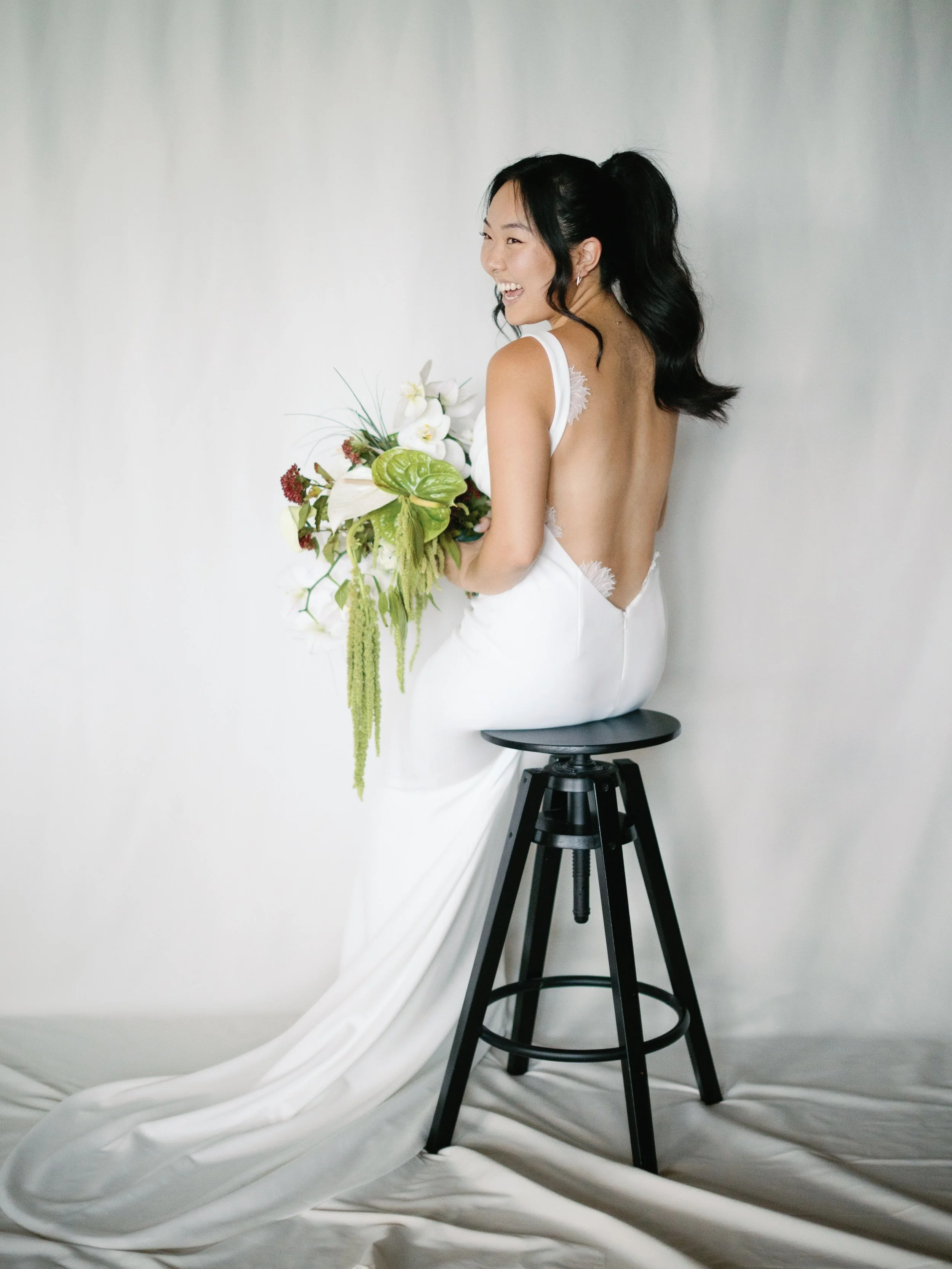 A woman in a white wedding dress sitting on a black stool, holding a bouquet of white and green flowers, smiling and looking to her left against a plain light background.