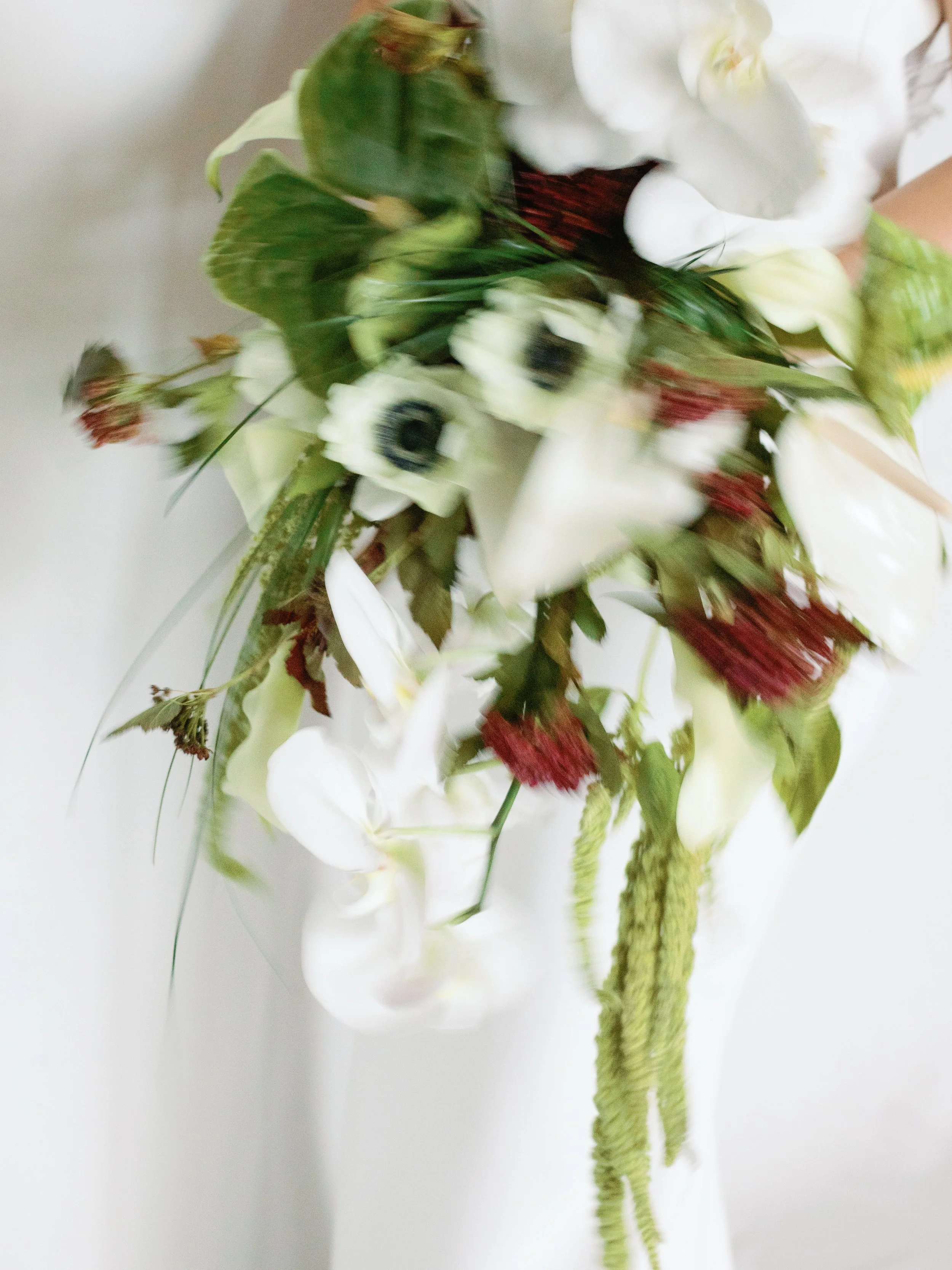 A blurry bouquet of white flowers, dark anemones, green leaves, and red accents being held in a person's hand.