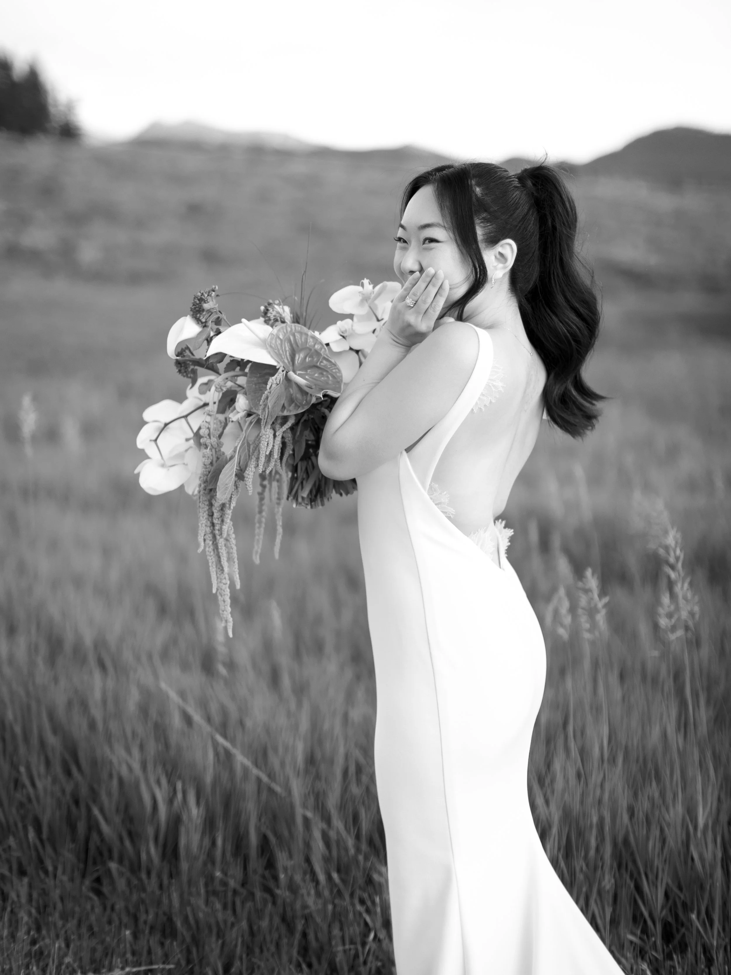 A woman in a wedding dress holding a large bouquet of flowers in an outdoor setting, smiling and covering her mouth with her hand.
