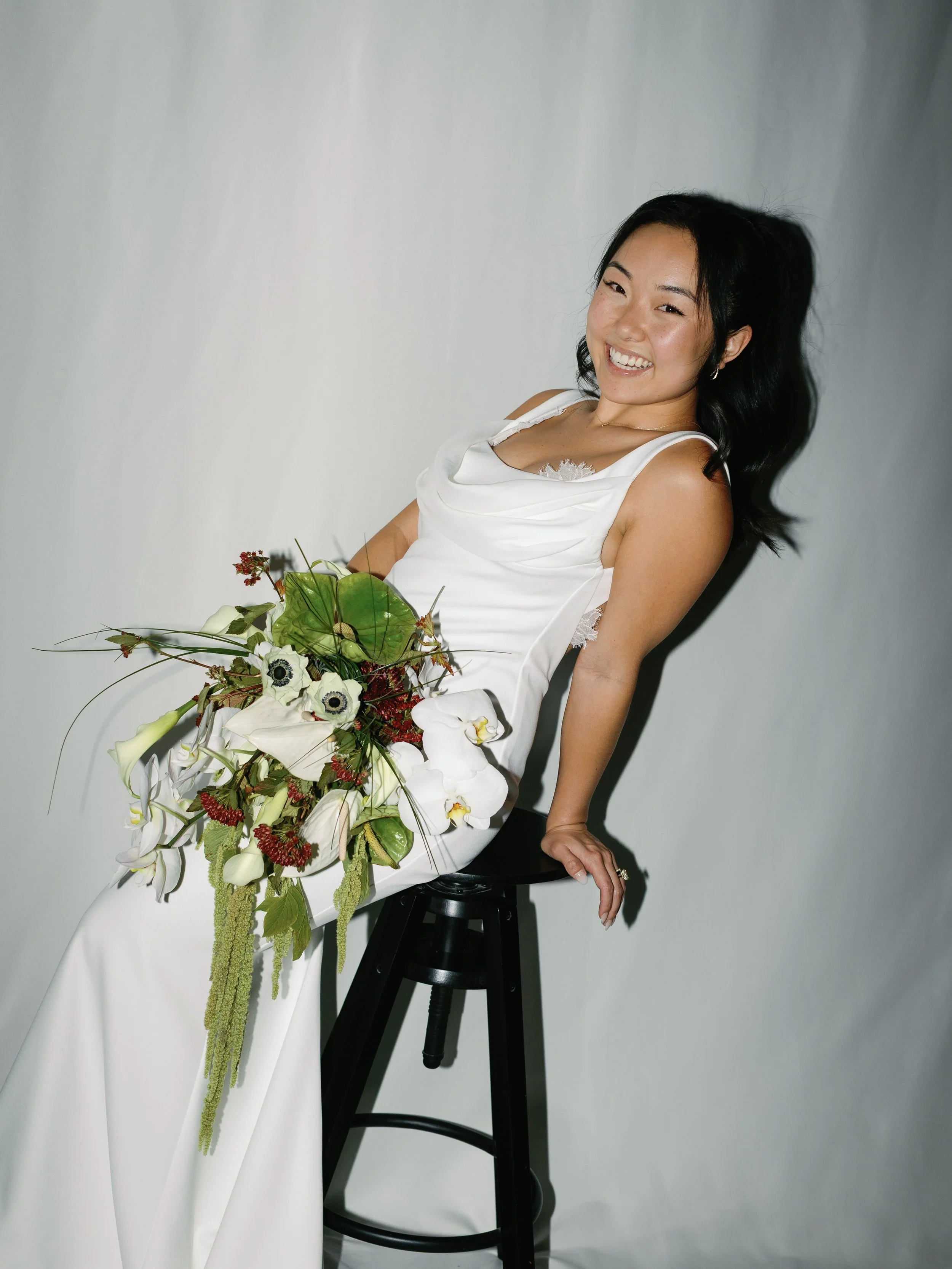 A smiling woman in a white dress holding a bouquet of white and green flowers, sitting on a black stool against a plain background.