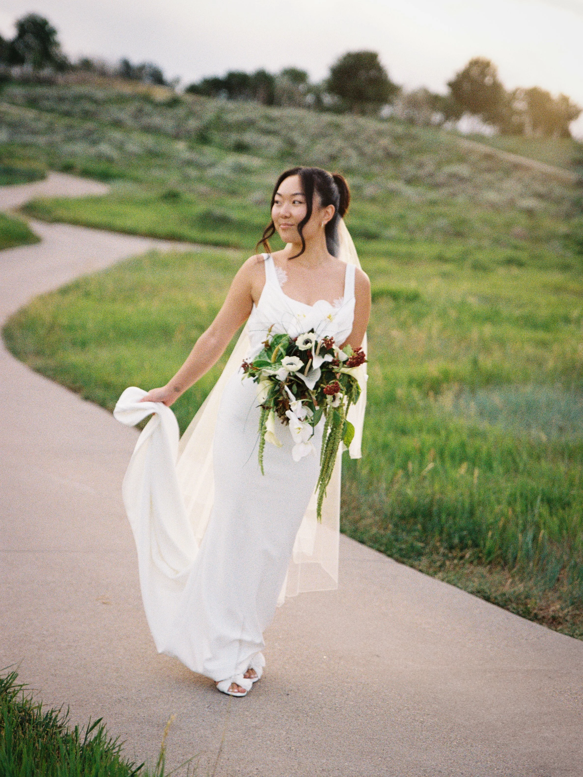 A bride in a white wedding dress holding a large bouquet of flowers, walking on a curved pathway surrounded by green grass and trees during sunset.