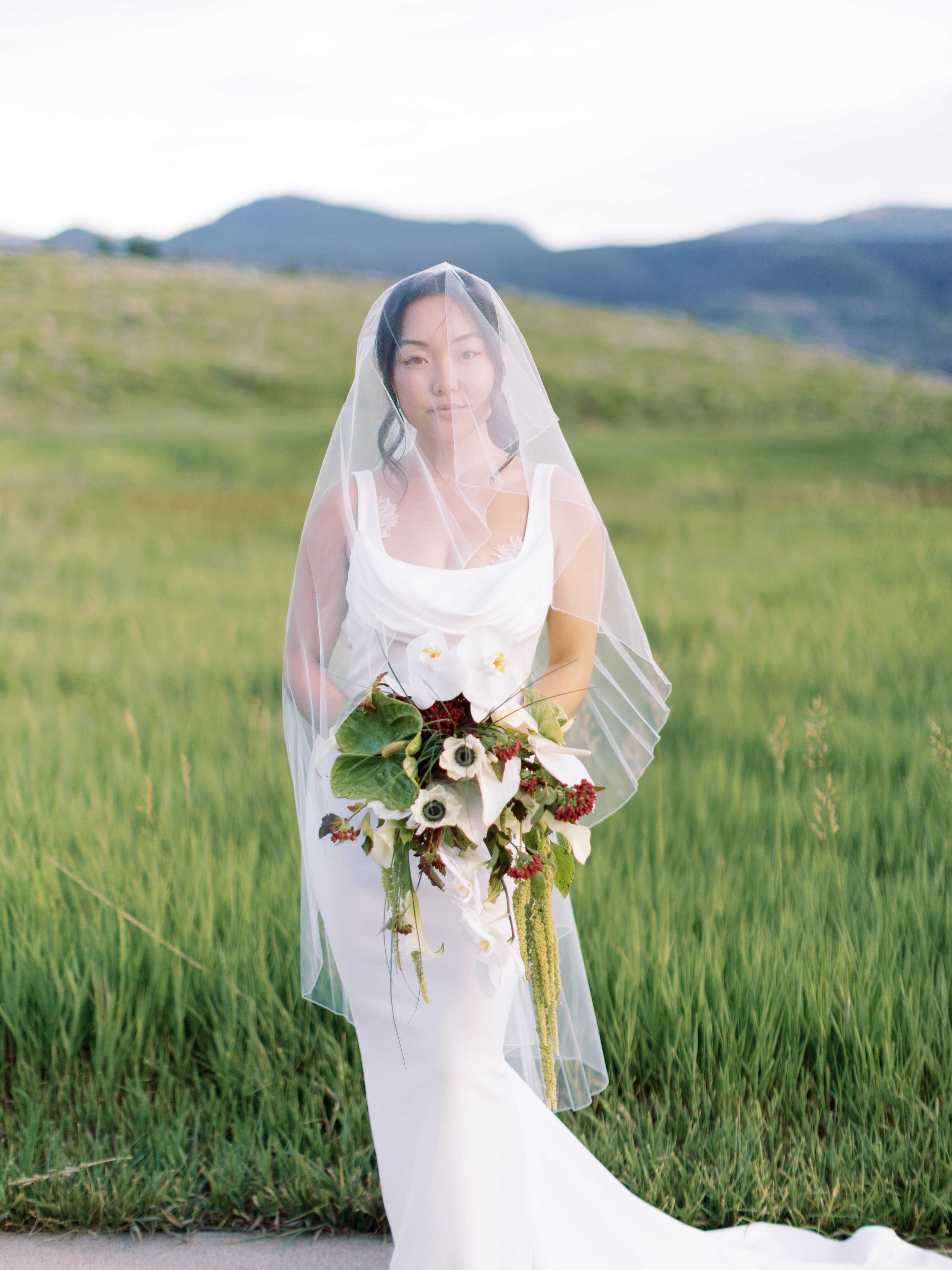 A woman in a white wedding dress with a veil holding a bouquet of white and green flowers standing in a green field with mountains in the background.