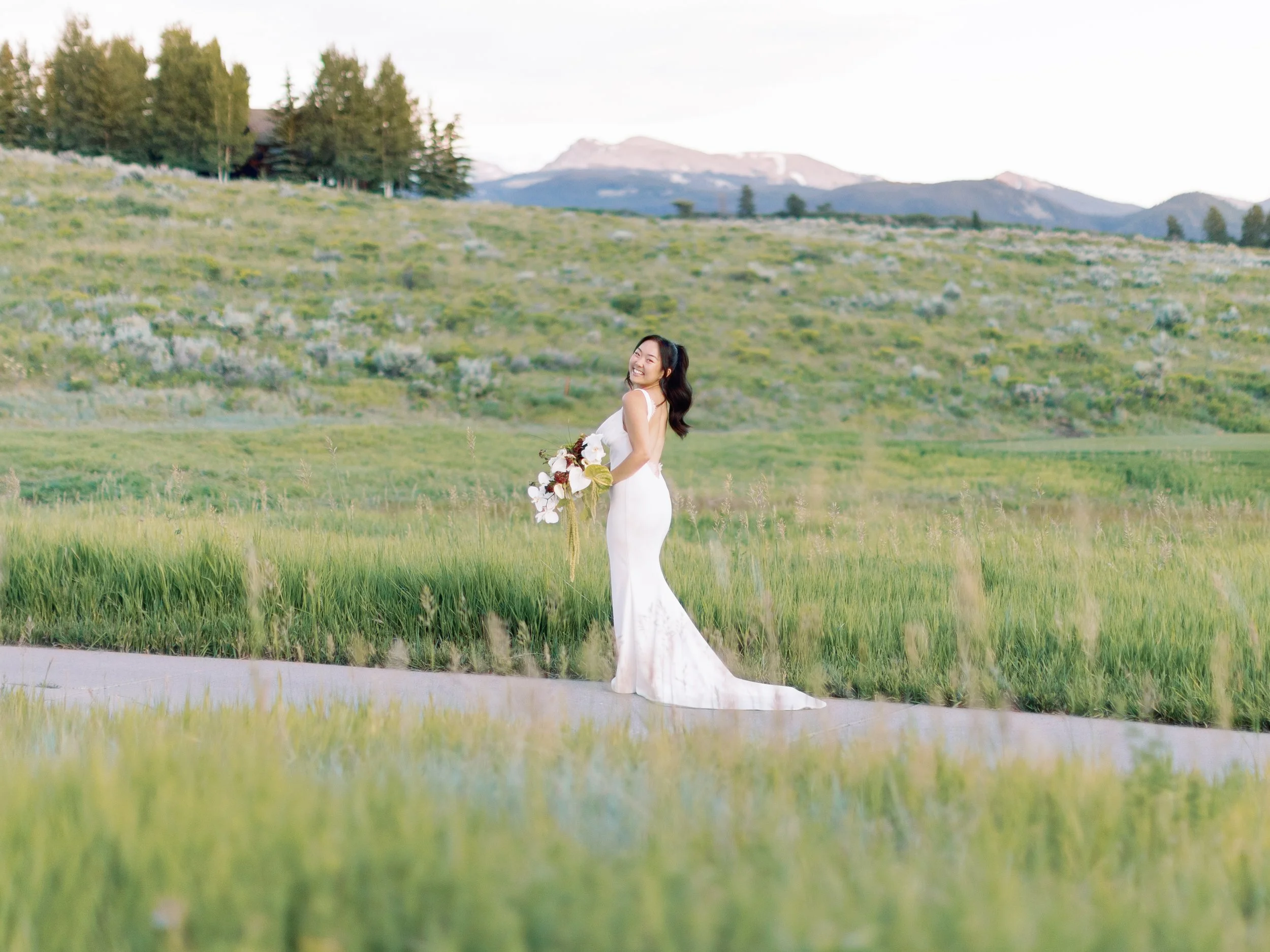 A woman in a white wedding dress holding a bouquet, standing on a grassy path in a green field with mountains in the background.