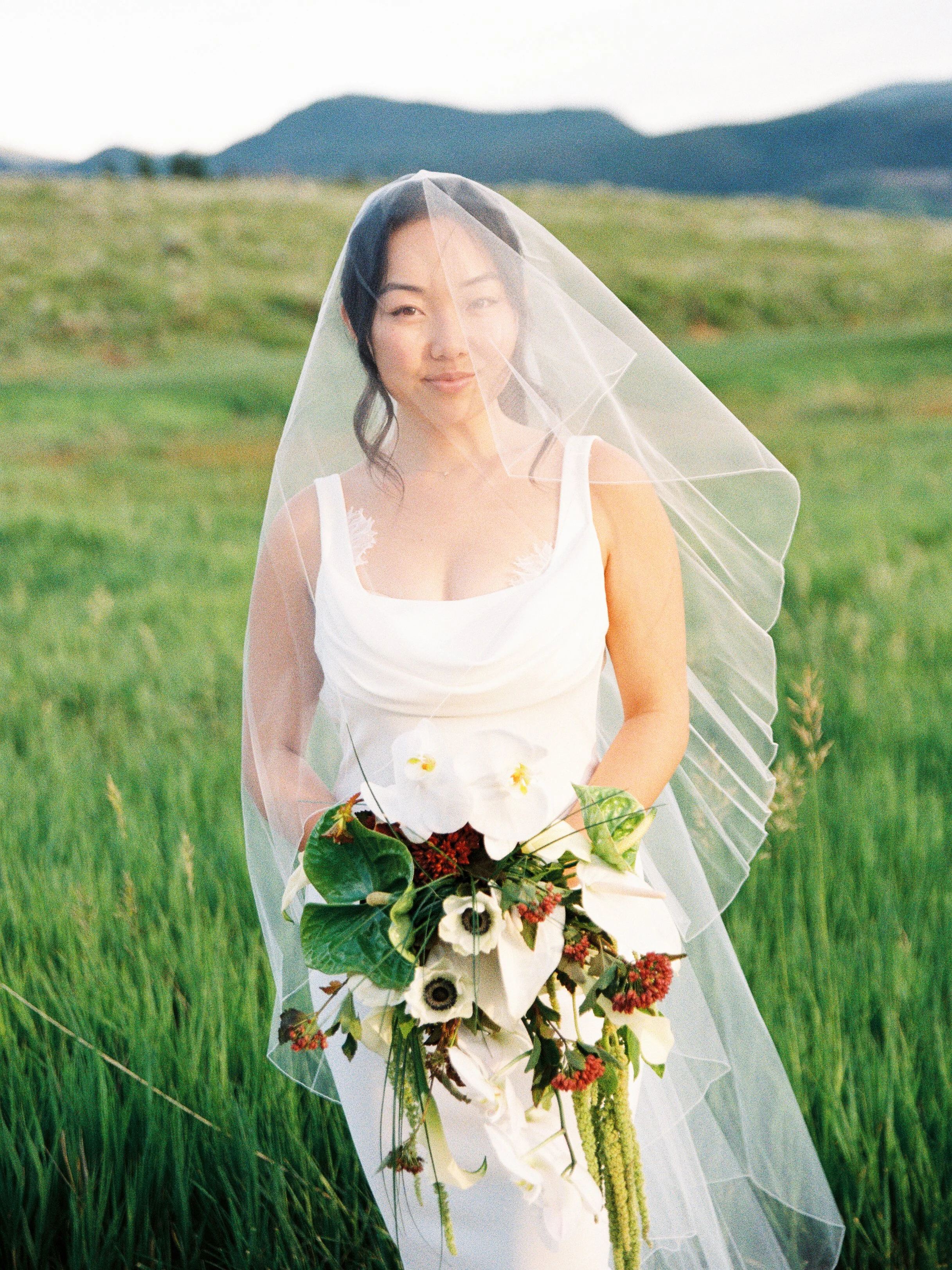 Bride in a white gown holding a bouquet of white calla lilies and greenery, standing in a grassy field with mountains in the background, wearing a sheer veil.