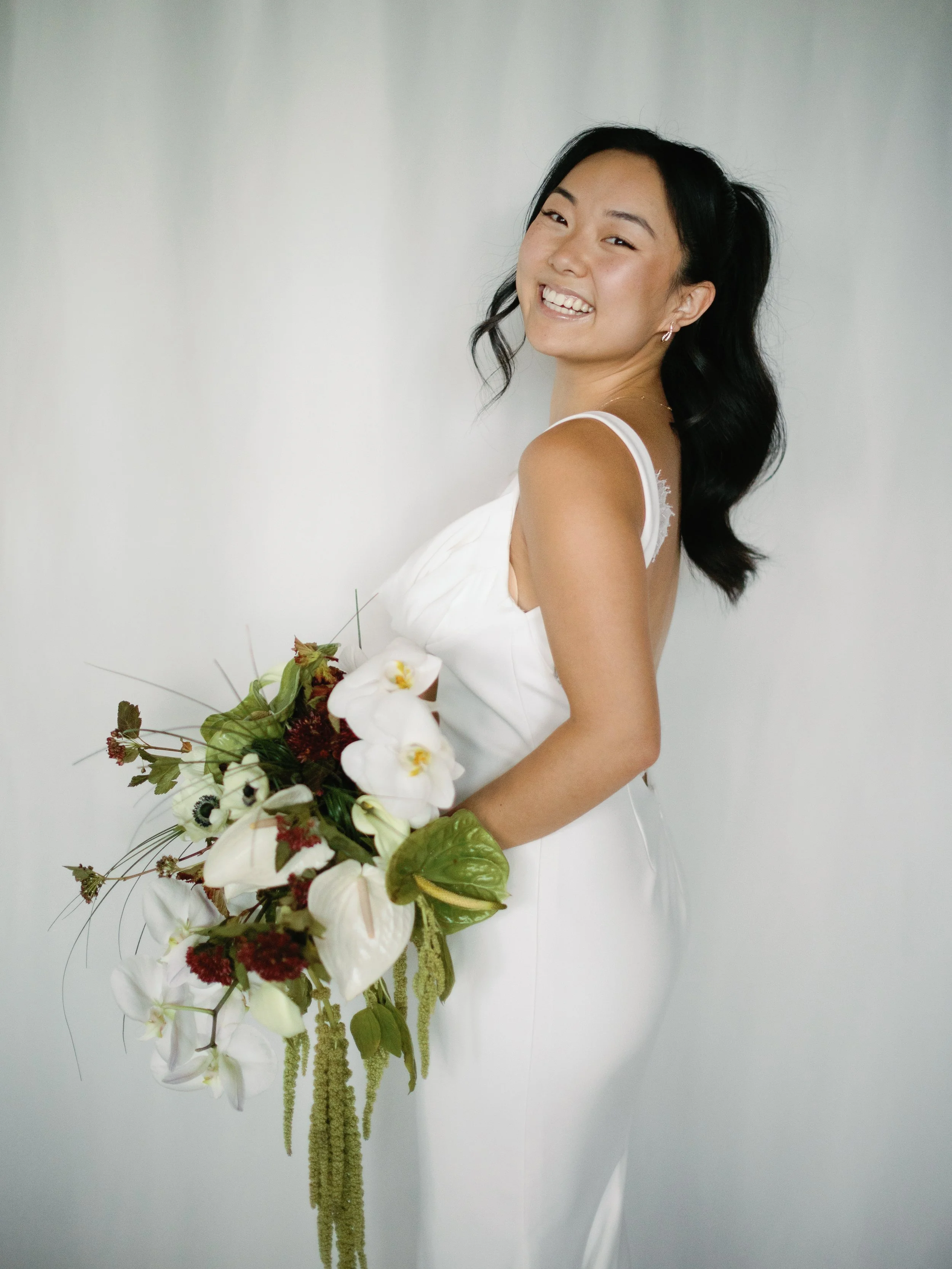 A woman in a white dress holding a bouquet of white flowers, smiling and looking at the camera.