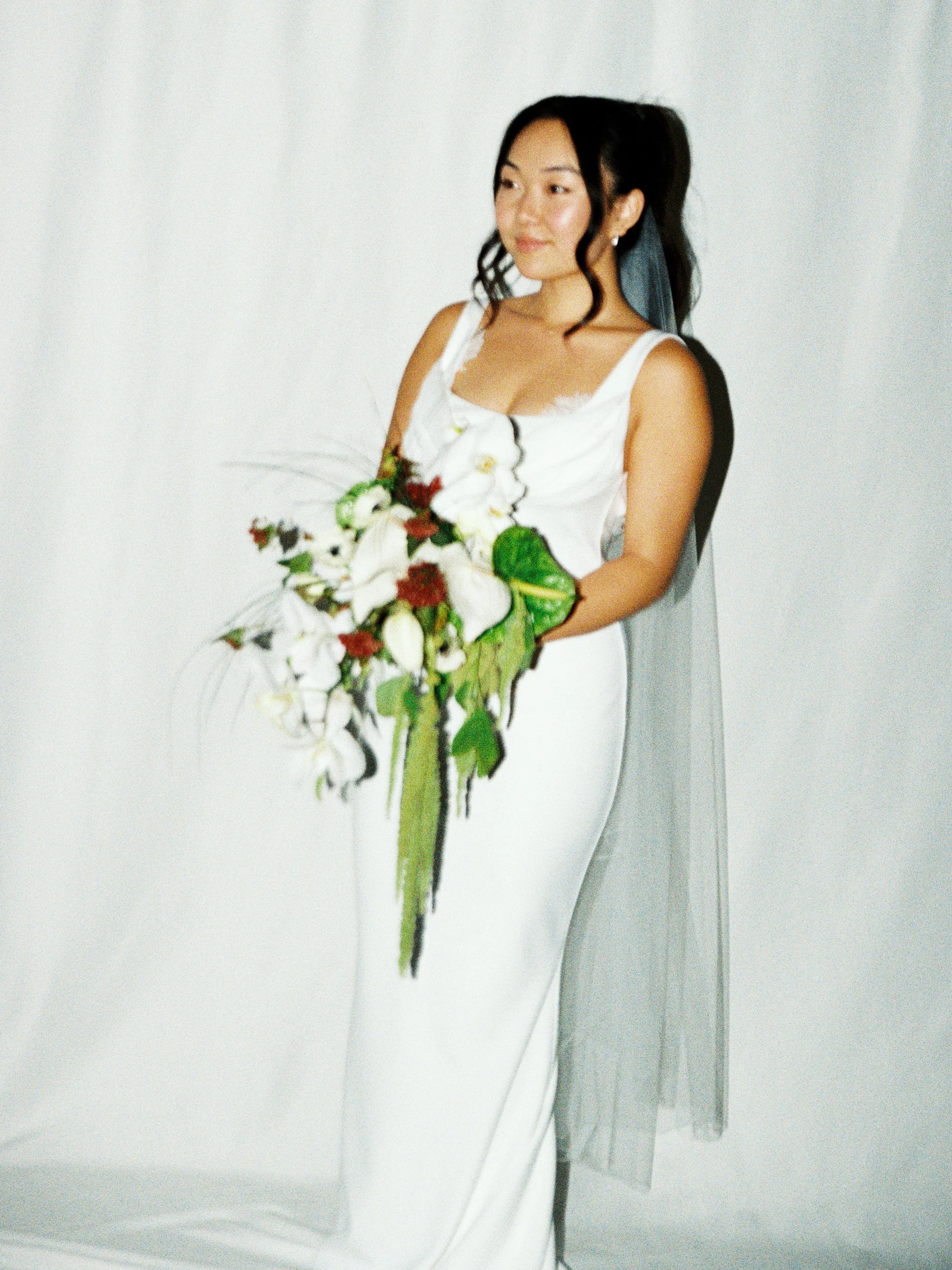 A woman in a white dress holding a bouquet of white and red flowers, standing in front of a plain white background.