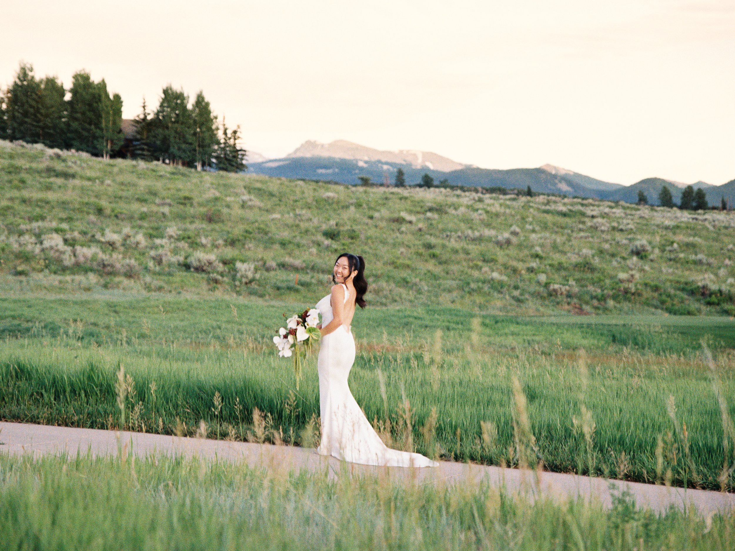 A woman in a white wedding dress holding a large bouquet of flowers, standing on a paved path in a lush green field with mountains and trees in the background during sunset.