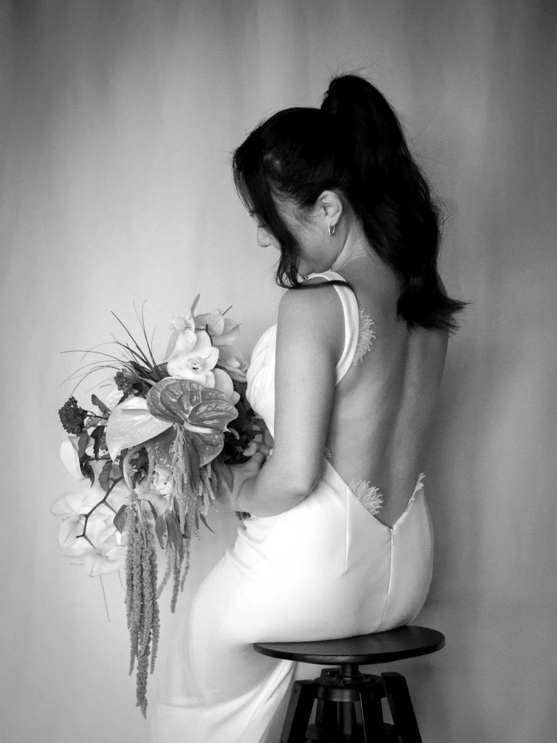 A woman in a backless white dress sits on a stool, holding a bouquet of flowers, with her head slightly tilted downward, dark hair styled in waves, photographed in black and white.