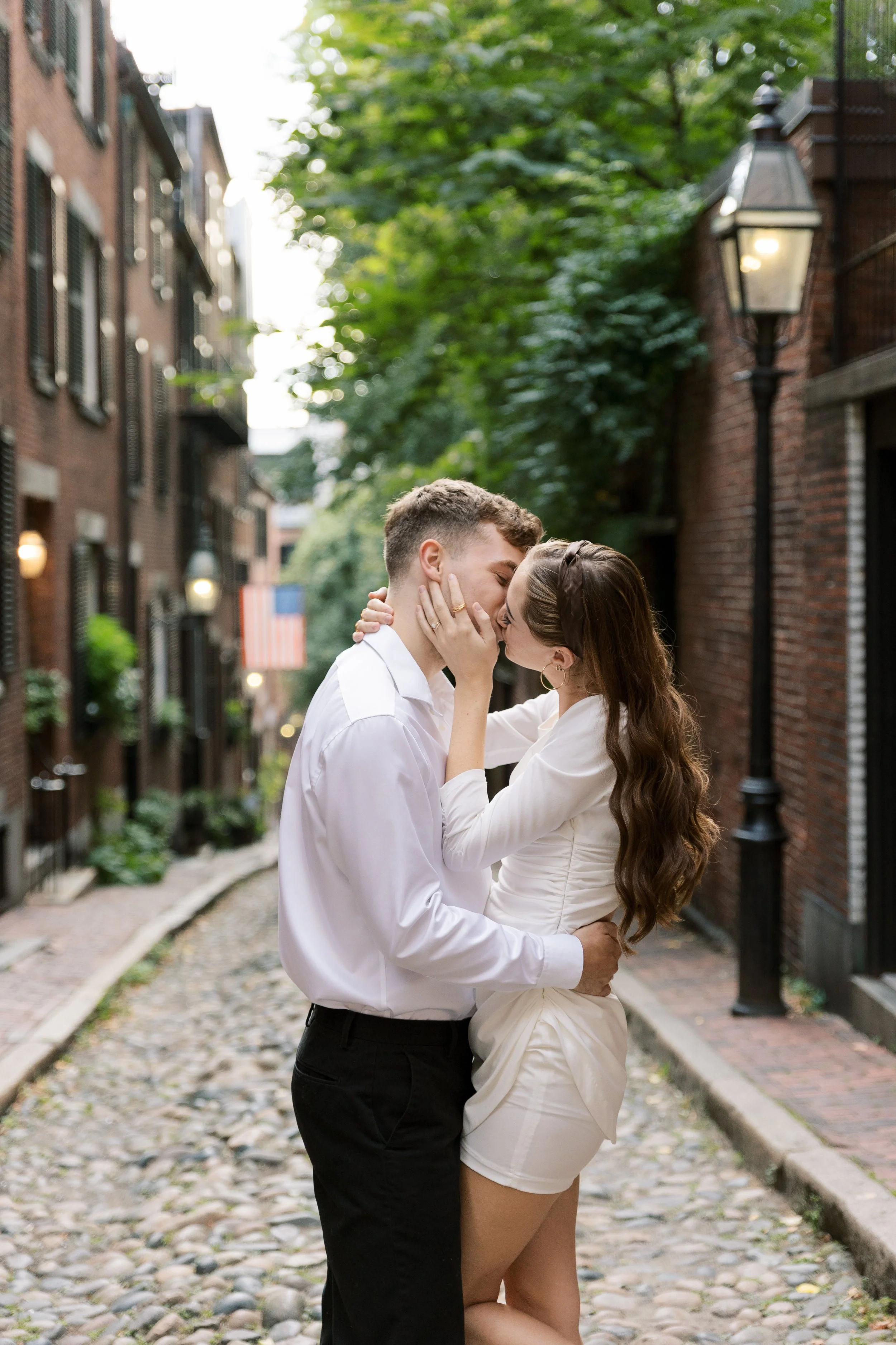 A young couple kissing on a cobblestone street in a city neighborhood during daytime, surrounded by brick buildings and green trees.