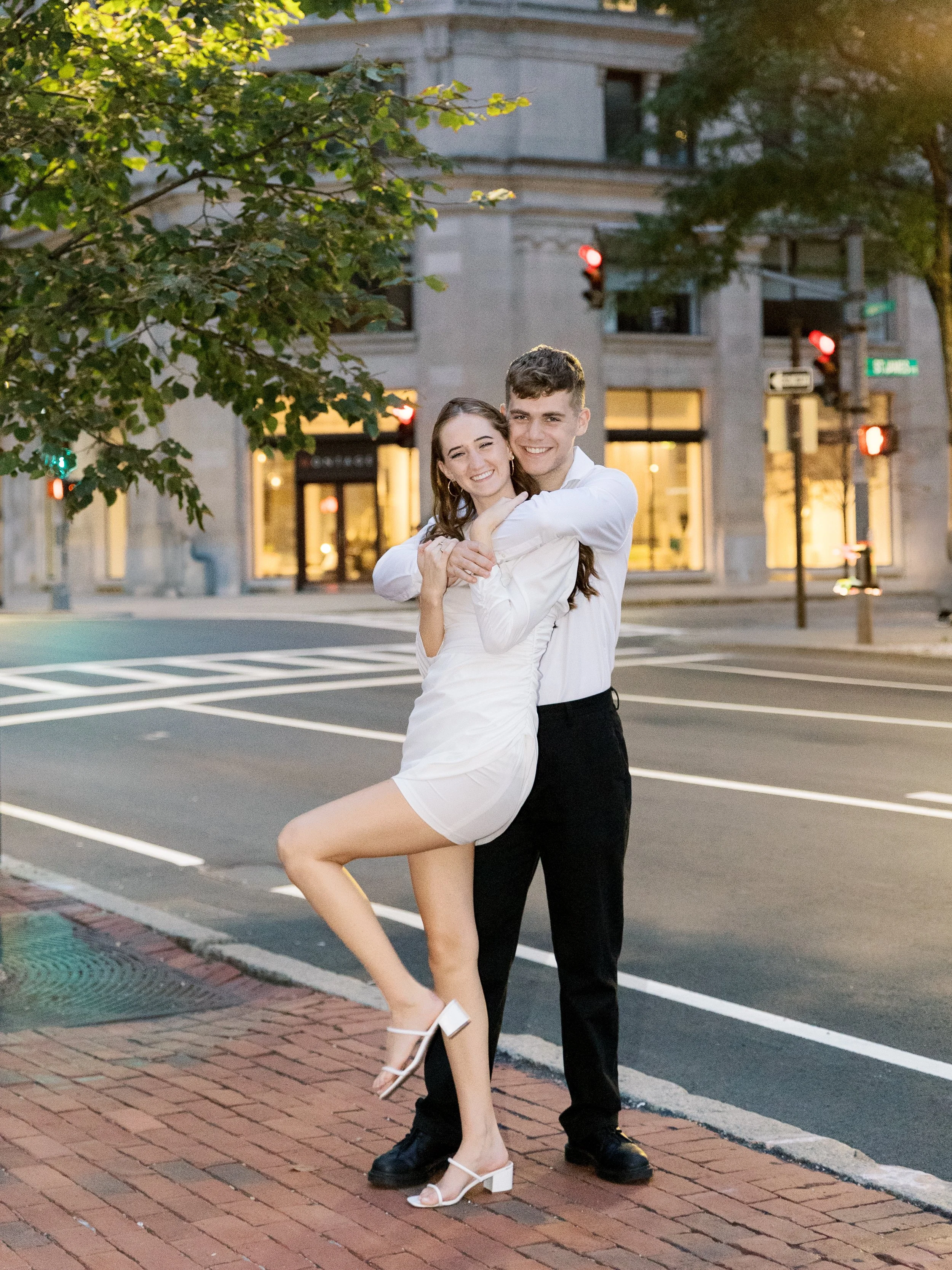 A young couple dressed in white, happily hugging and smiling on a city street during evening time.