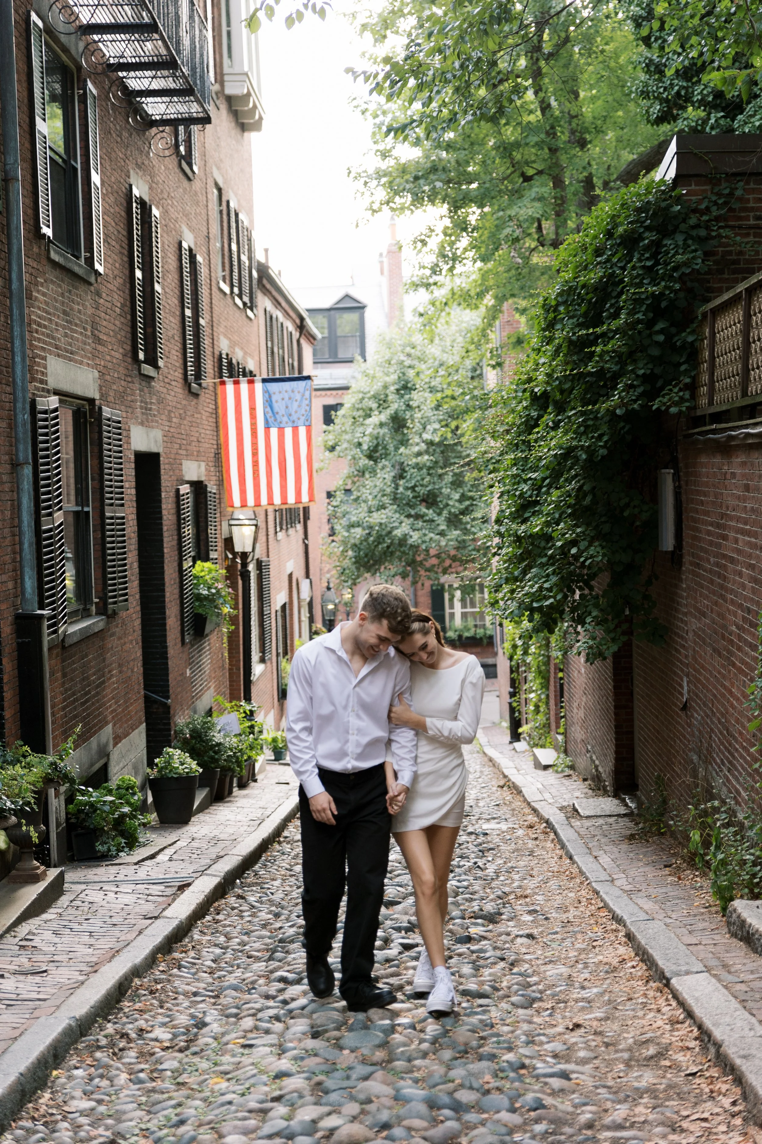 A young couple holding hands and walking together on a cobblestone street in an urban neighborhood, with brick buildings, greenery, and an American flag hanging on a building.